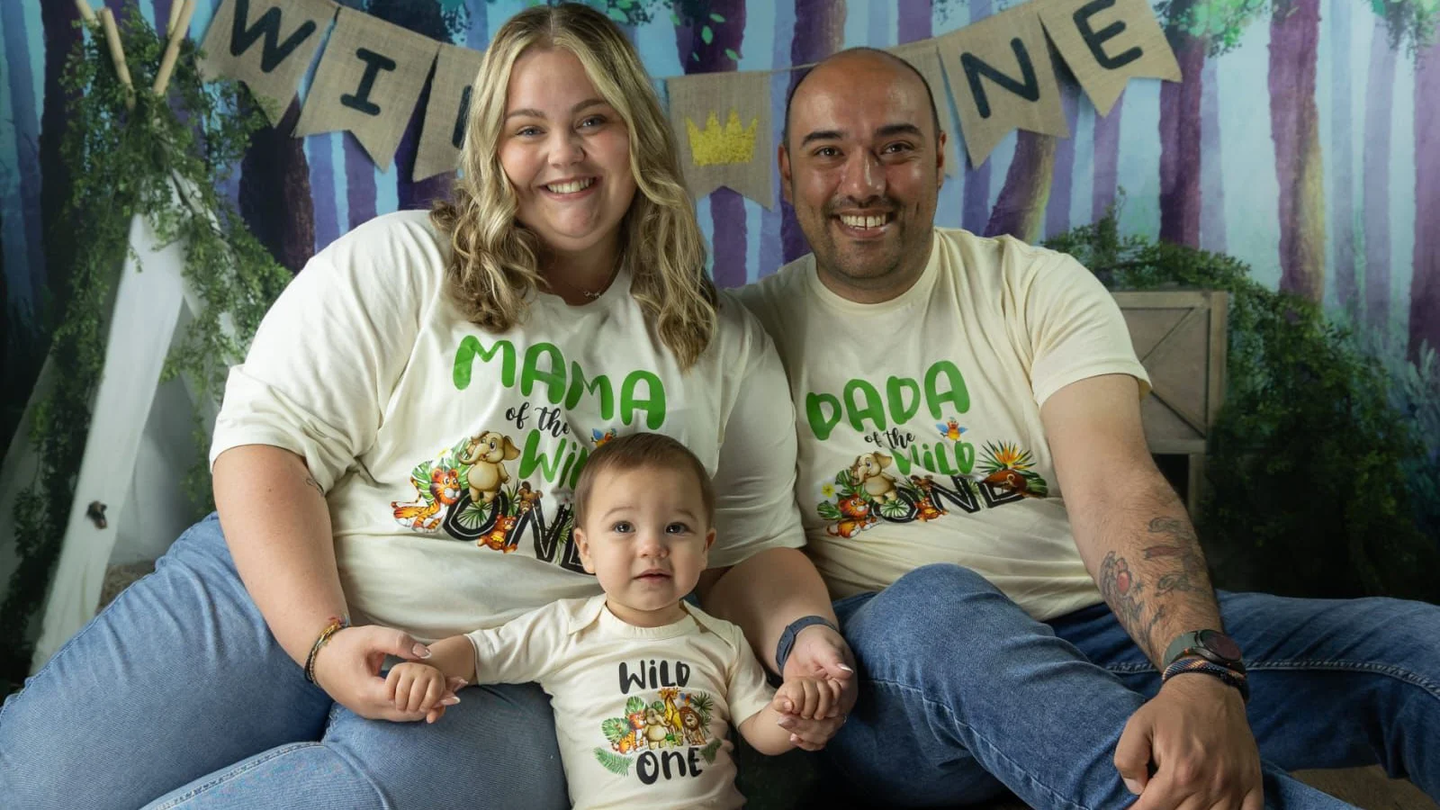 A photograph of Leah Weenen, her baby, Cayden, and husband Charlie Abel, smiling to camera and wearing matching 'Mama' and 'Dada' t-shirts.