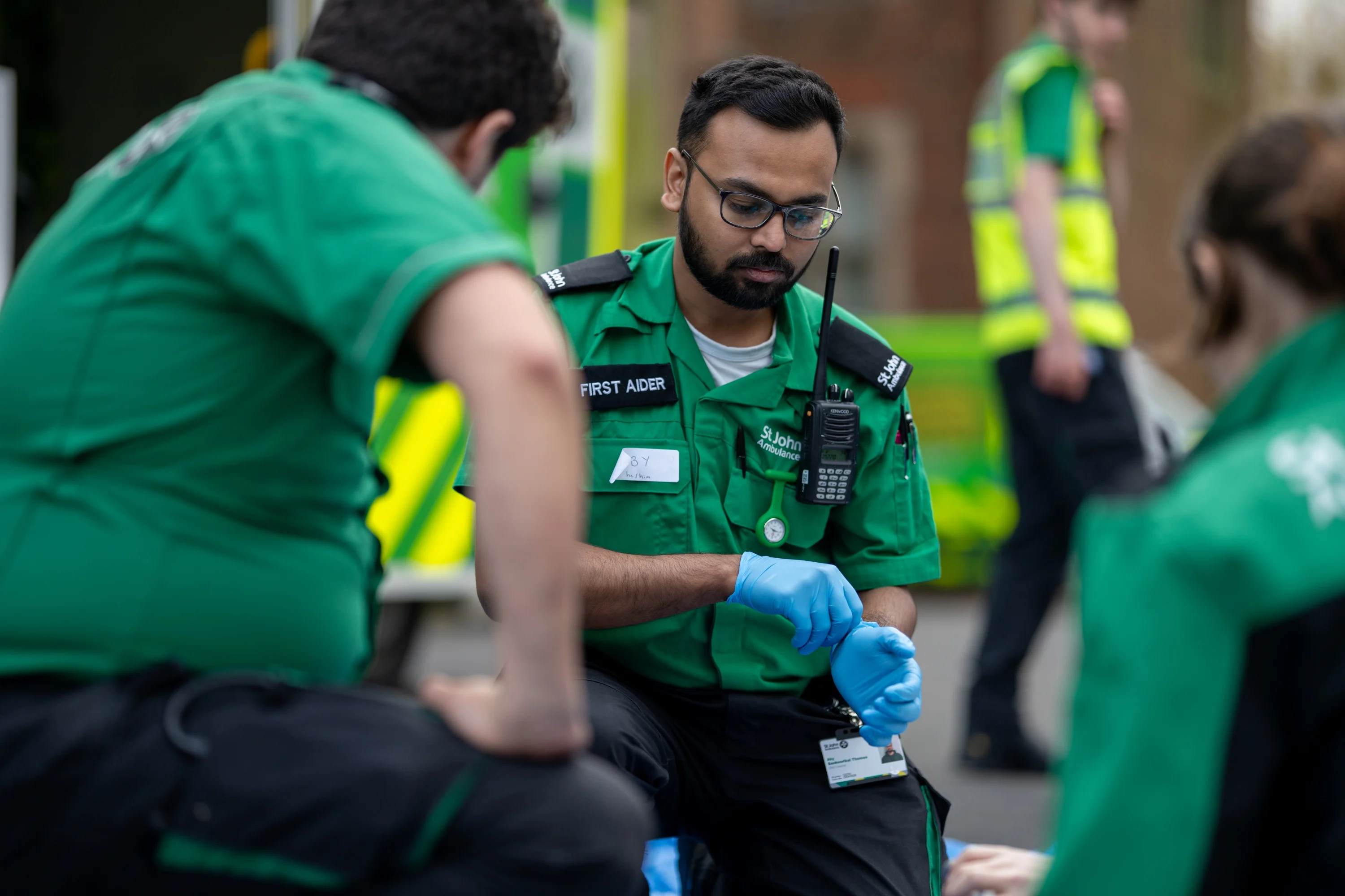 A First Aider kneeling on the ground looking at something on the floor. They are putting on blue medical gloves. There are two other First Aiders kneeling in front of them. In the background, there is an ambulance and another person standing wearing a high-vis vest.