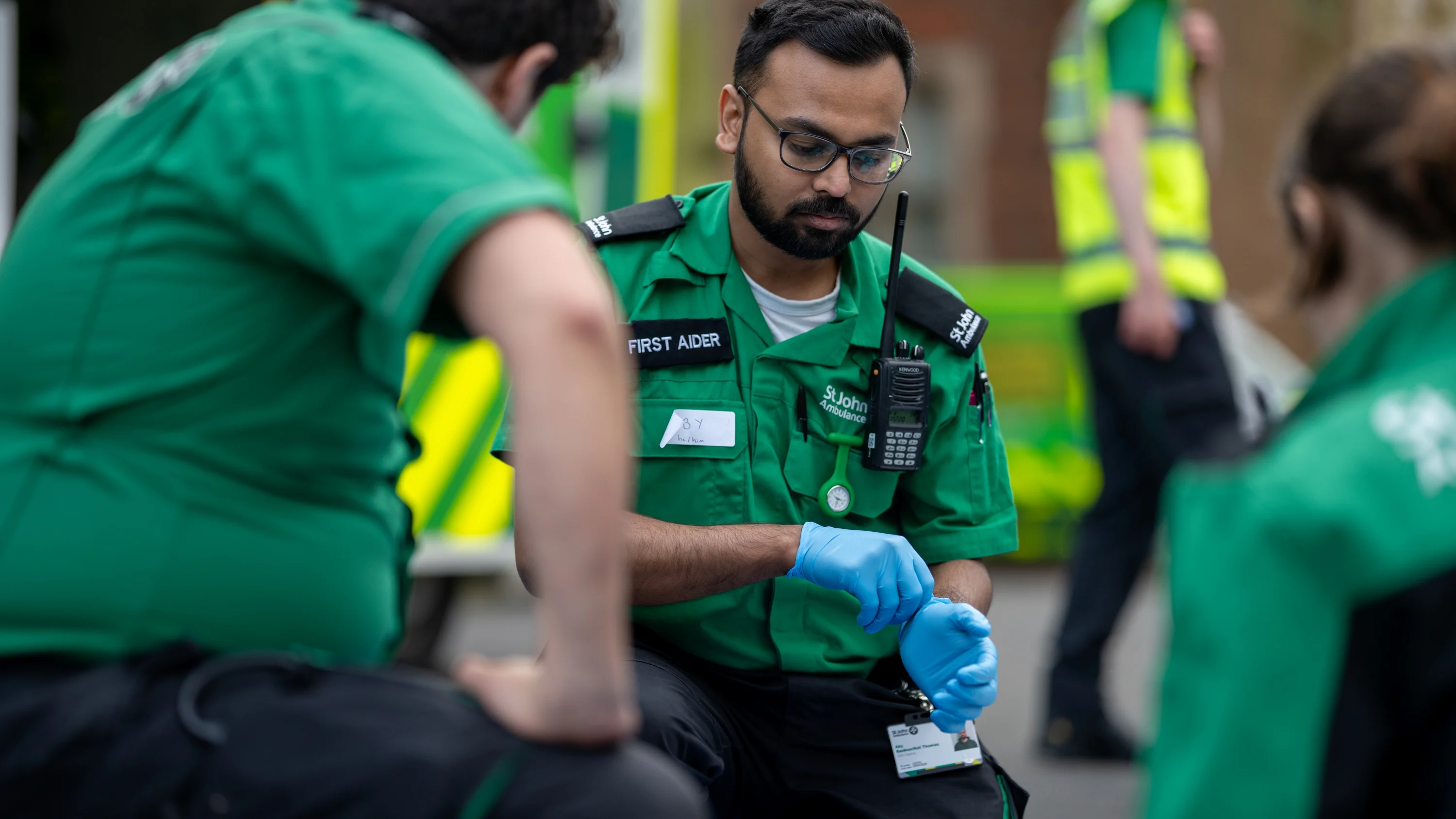 A First Aider kneeling on the ground looking at something on the floor. They are putting on blue medical gloves. There are two other First Aiders kneeling in front of them. In the background, there is an ambulance and another person standing wearing a high-vis vest.