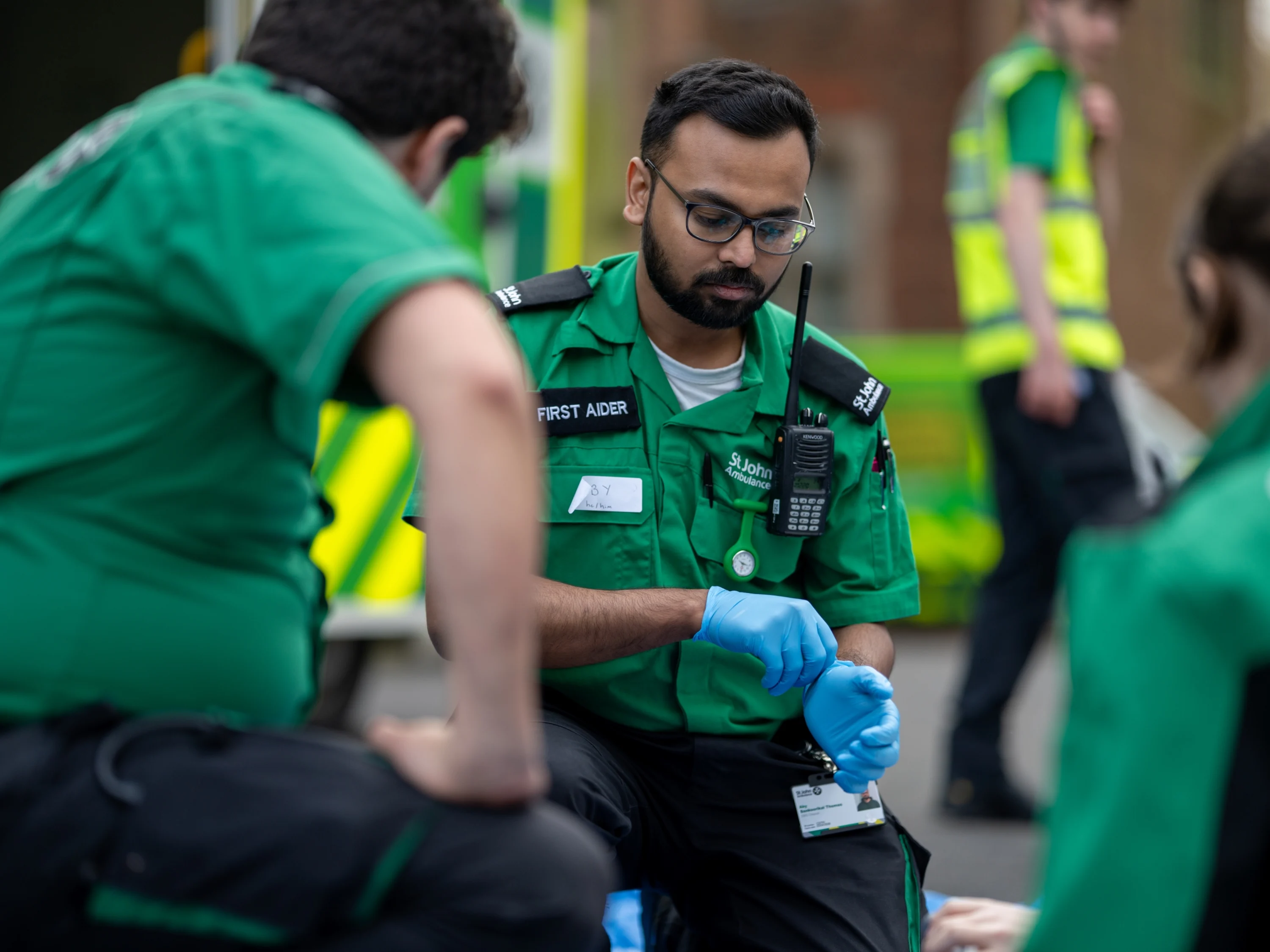 A First Aider kneeling on the ground looking at something on the floor. They are putting on blue medical gloves. There are two other First Aiders kneeling in front of them. In the background, there is an ambulance and another person standing wearing a high-vis vest.