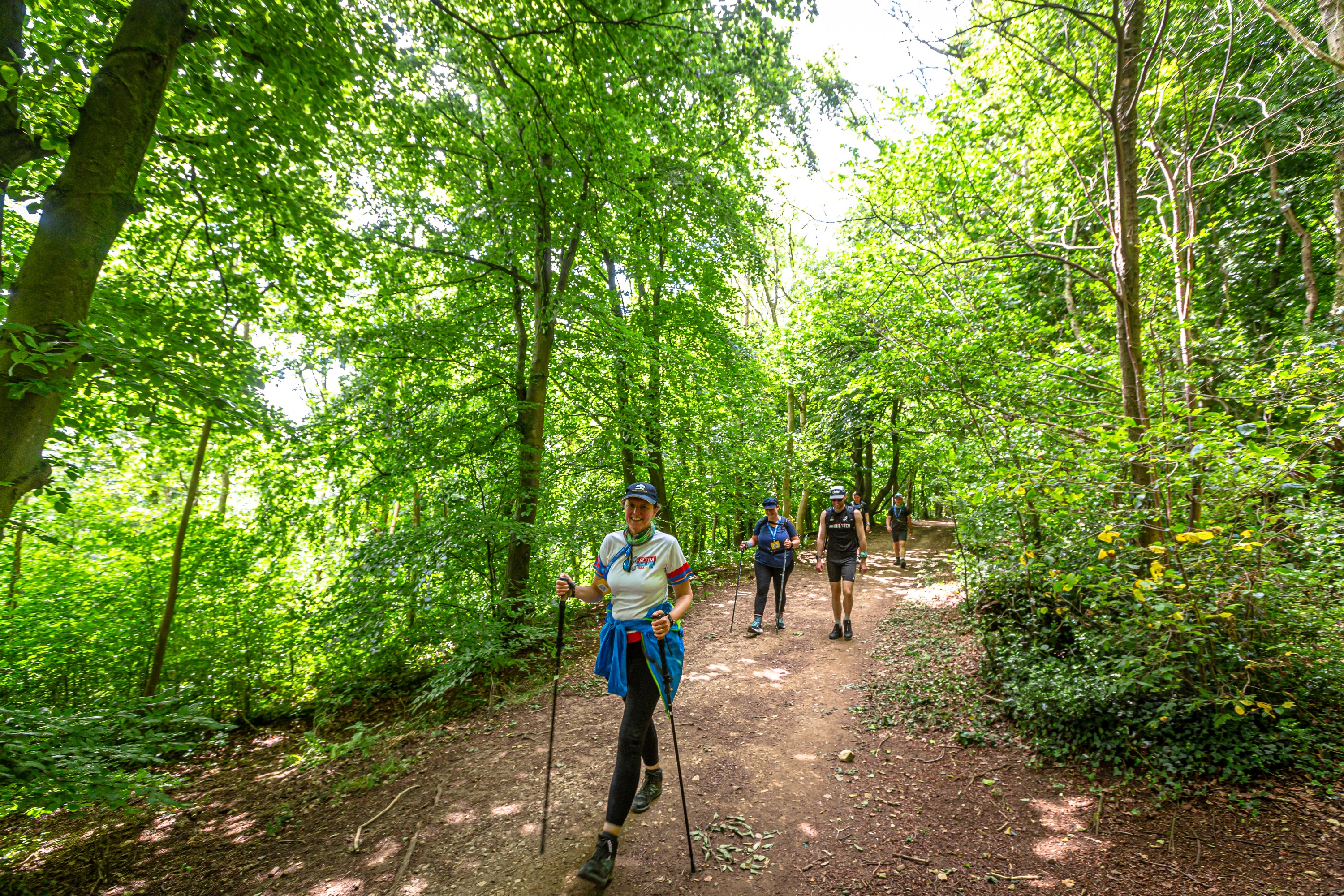 Three people with walking sticks walking along a footpath through some woods. 