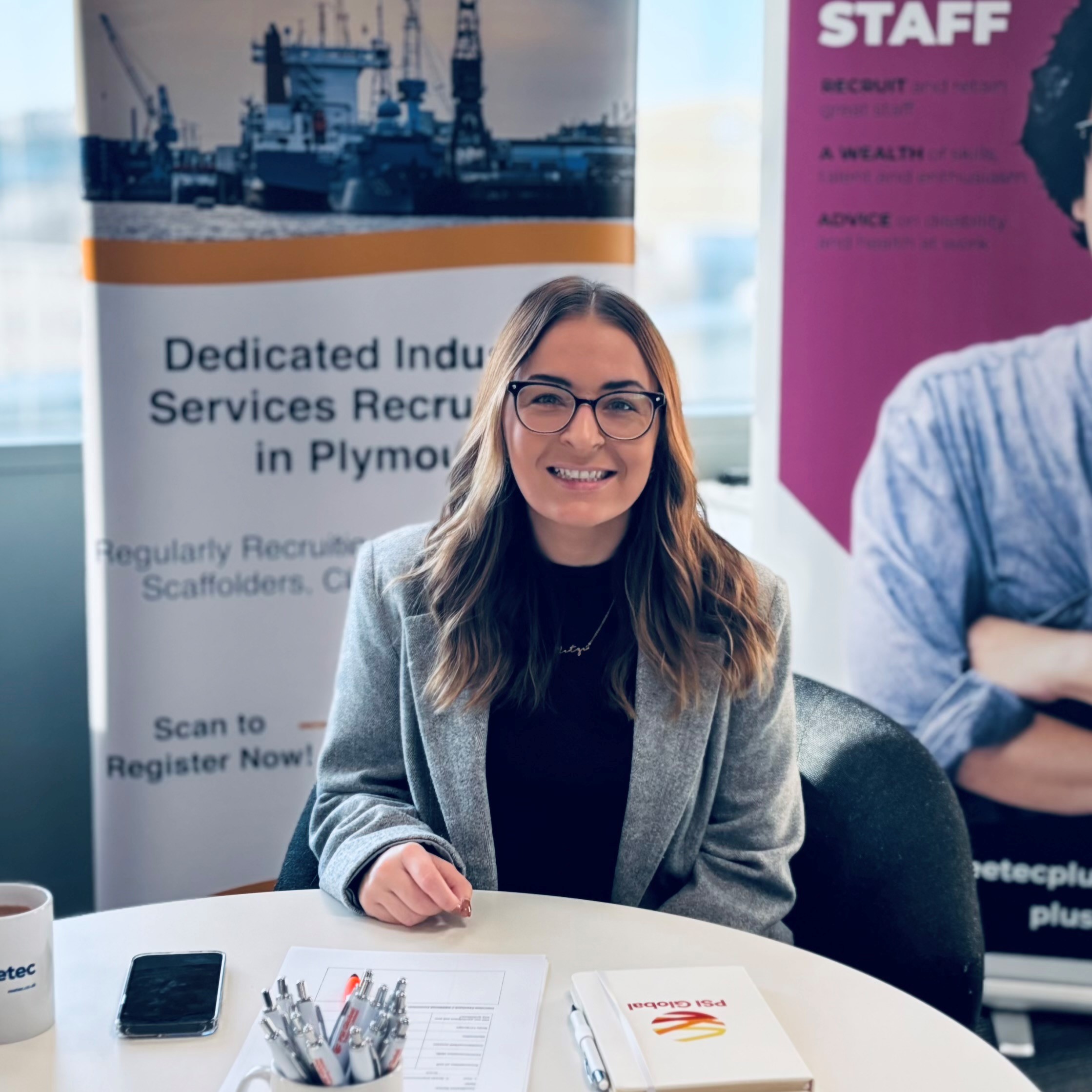 A photograph of Yasmin at a work event. She is sitting at a table and smiling to camera. In the background are banners advertising recruitment.