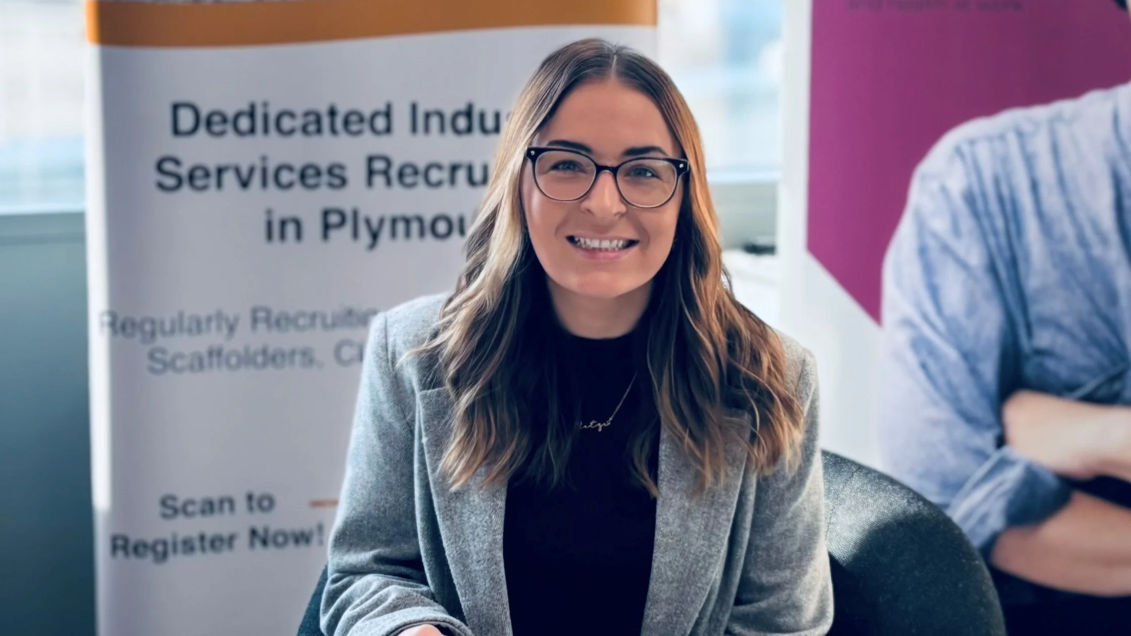 A photograph of Yasmin at a work event. She is sitting at a table and smiling to camera. In the background are banners advertising recruitment.