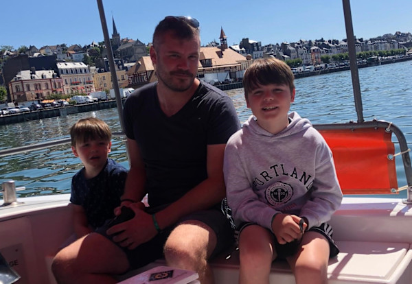 A photograph of James Berry and two of his sons sat on a boat on holiday and smiling towards the camera.