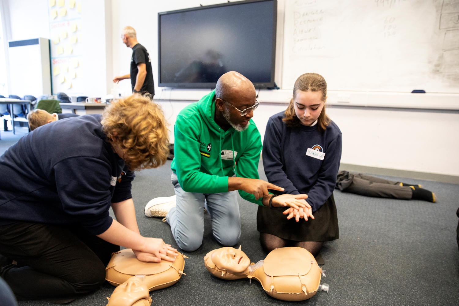 Three people in a classroom kneeling on the floor with CPR mannequins in front of them. One of them is showing a student how to position their hands for CPR. There are other people and CPR mannequins in the background. 