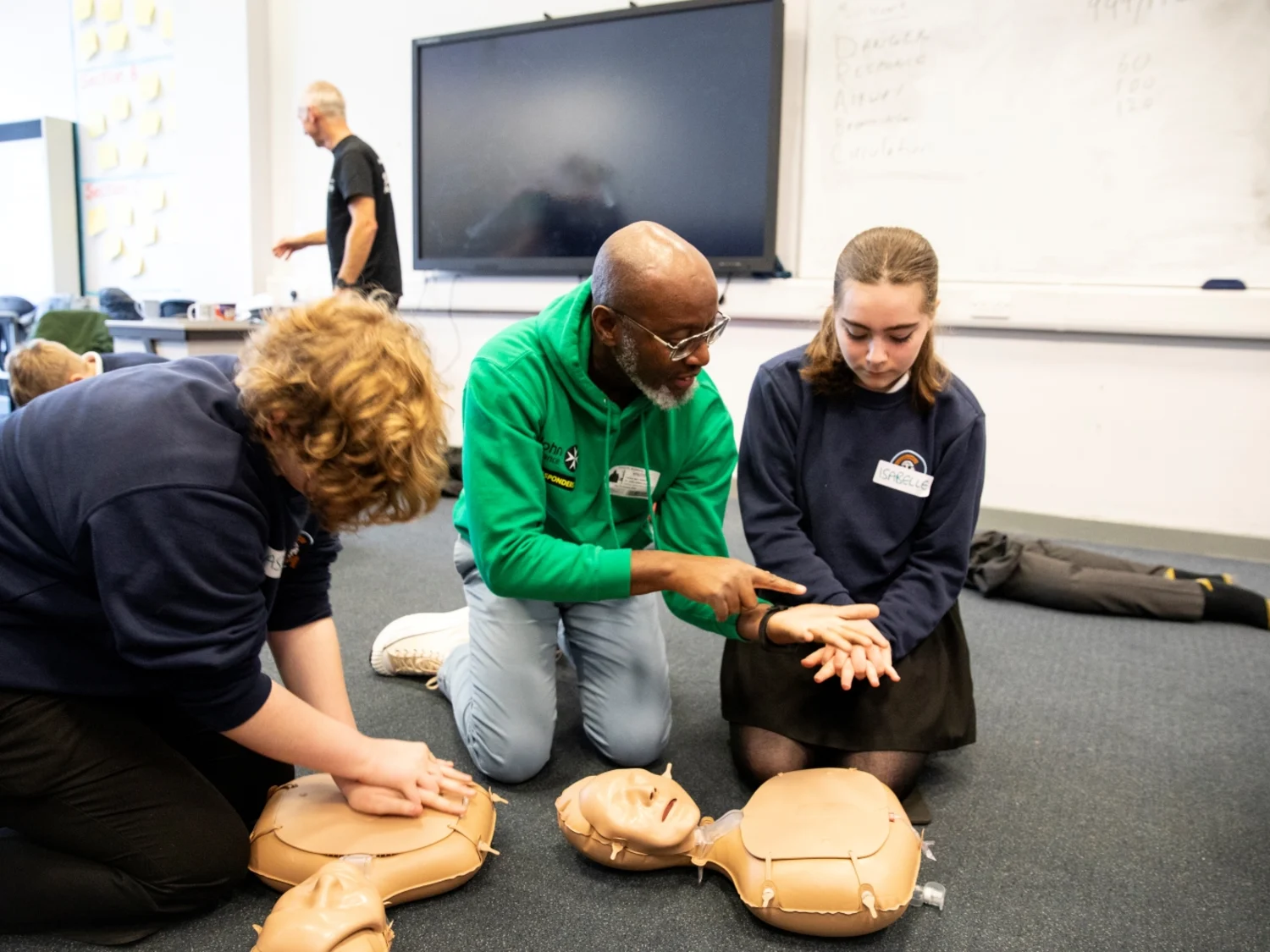 Three people in a classroom kneeling on the floor with CPR mannequins in front of them. One of them is showing a student how to position their hands for CPR. There are other people and CPR mannequins in the background.