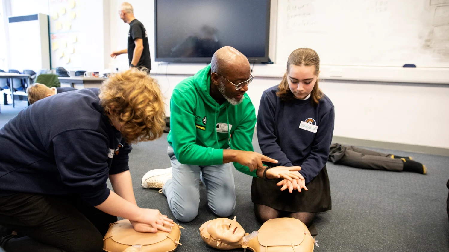 Three people in a classroom kneeling on the floor with CPR mannequins in front of them. One of them is showing a student how to position their hands for CPR. There are other people and CPR mannequins in the background.