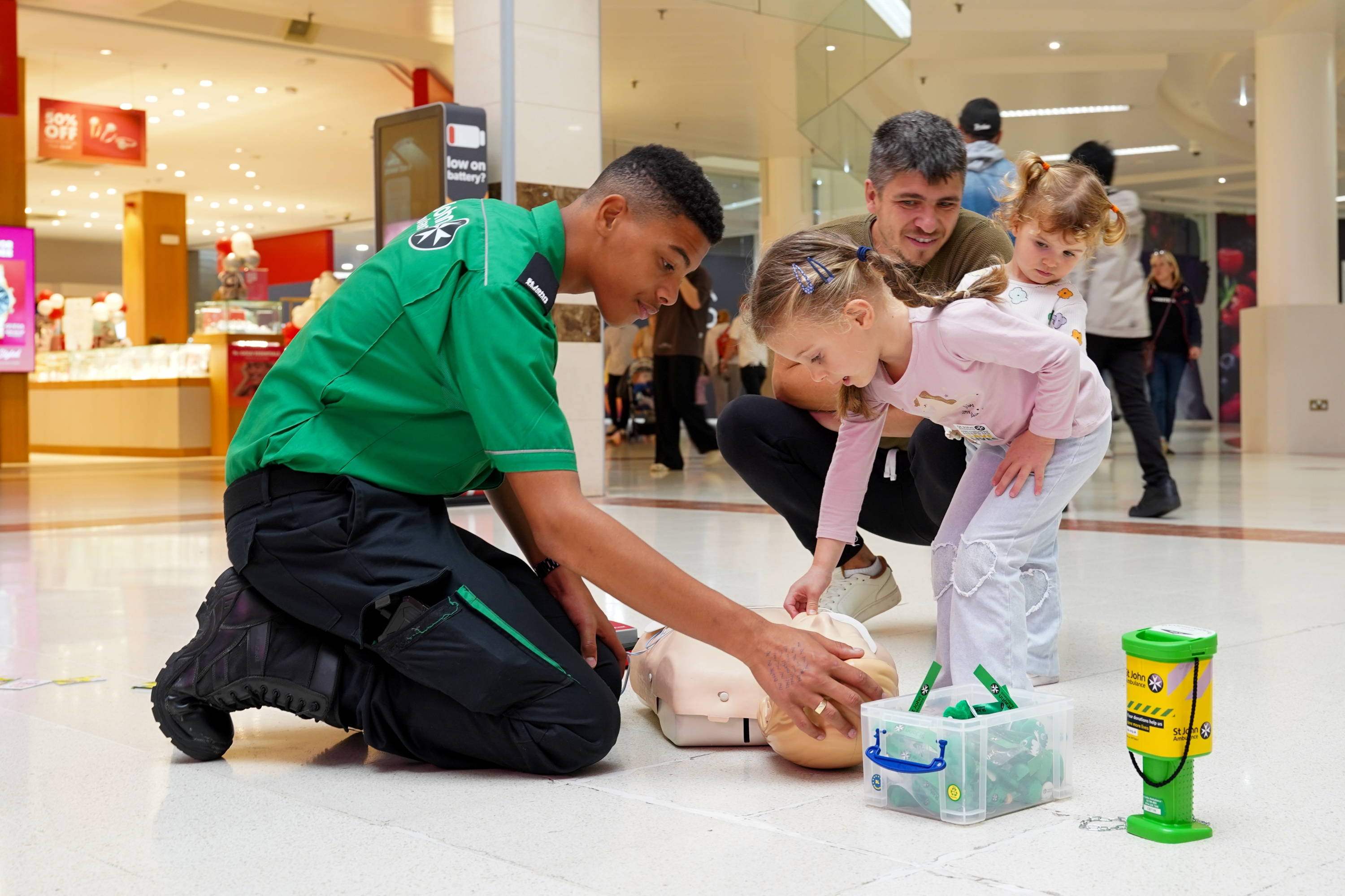 In a shopping centre, a St John Ambulance volunteer is kneeling down on the ground opposite a young child. Both are looking at and touching a CPR mannequin. 