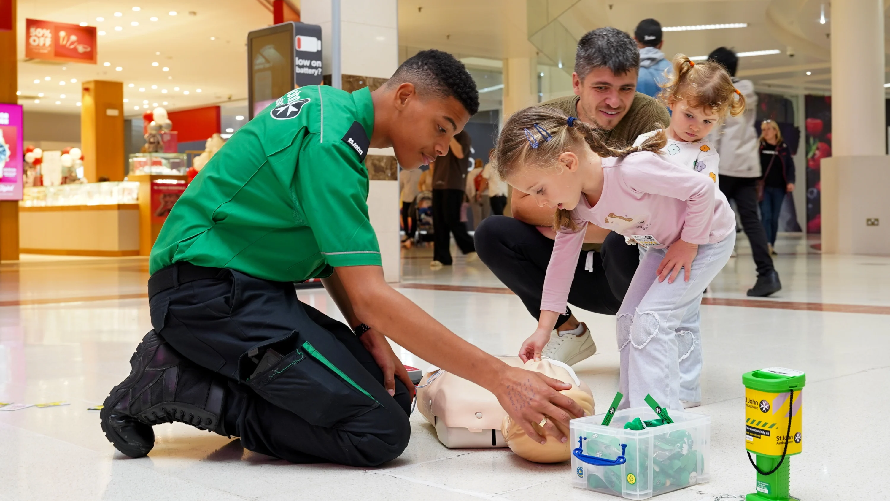 In a shopping centre, a St John Ambulance volunteer is kneeling down on the ground opposite a young child. Both are looking at and touching a CPR mannequin.