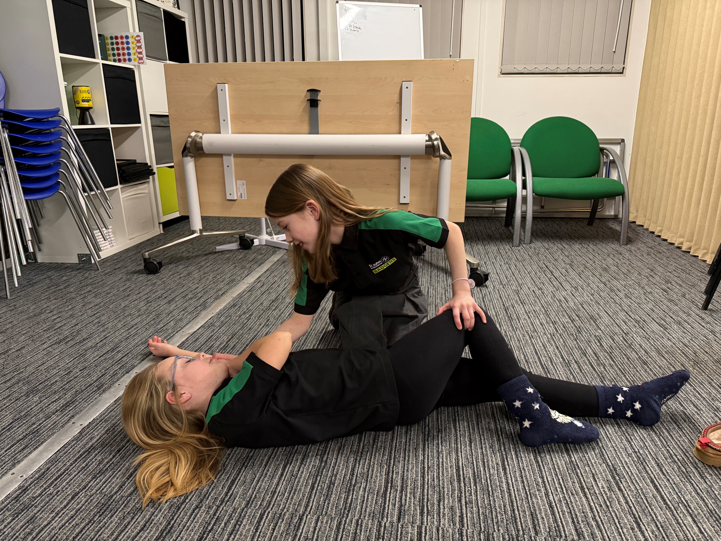 Two Badgers in a first aid training room. One is lying on the floor and the other is kneeling next to them putting them into the recovery position. 