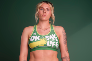 A photograph of Millie Bright wearing the SJA CPR Bra and standing in front of a dark green background. The CPR Bra reads: It's OK to save my life.