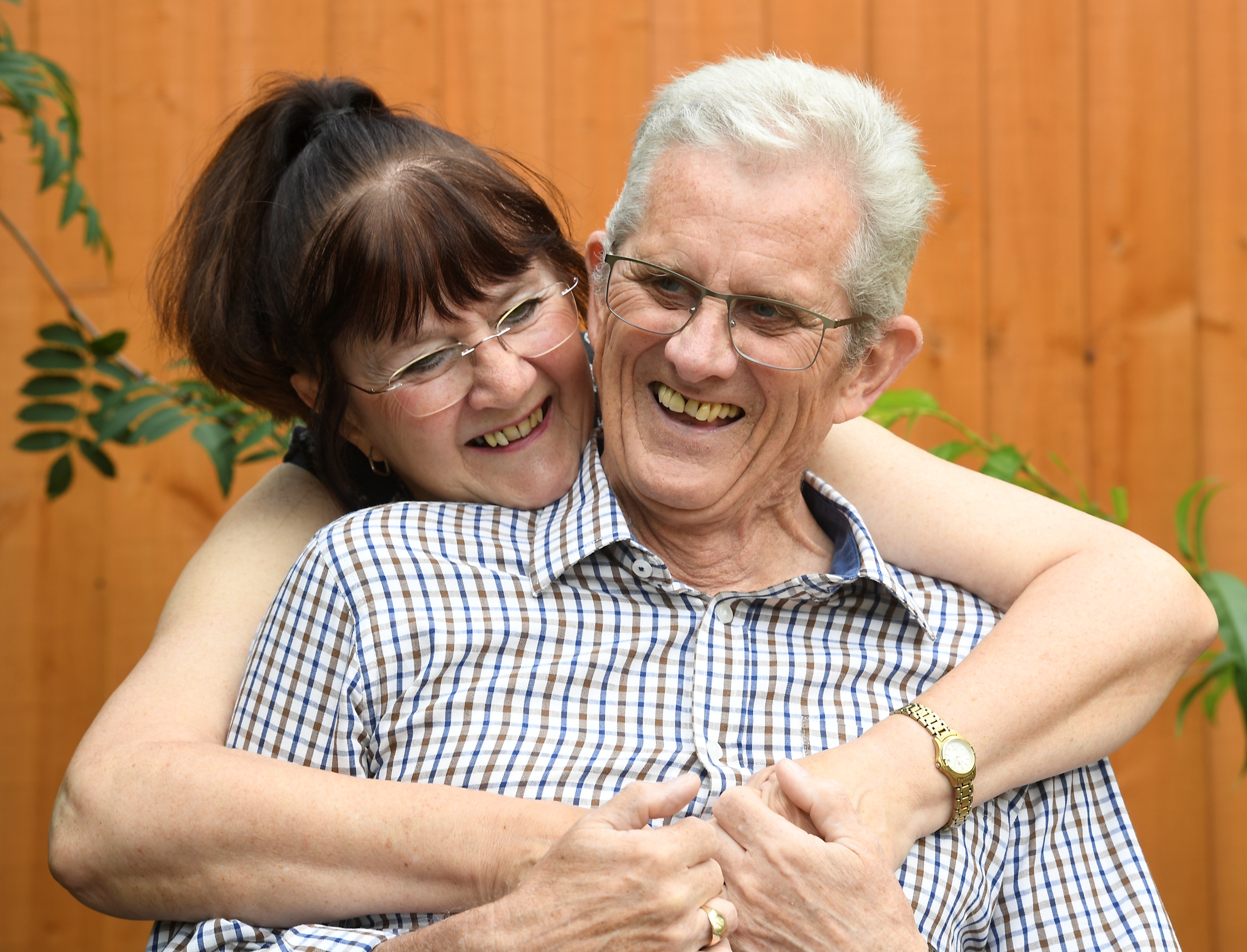 A photograph of Gwen and John in their garden. Gwen is hugging John from behind and they're both laughing.