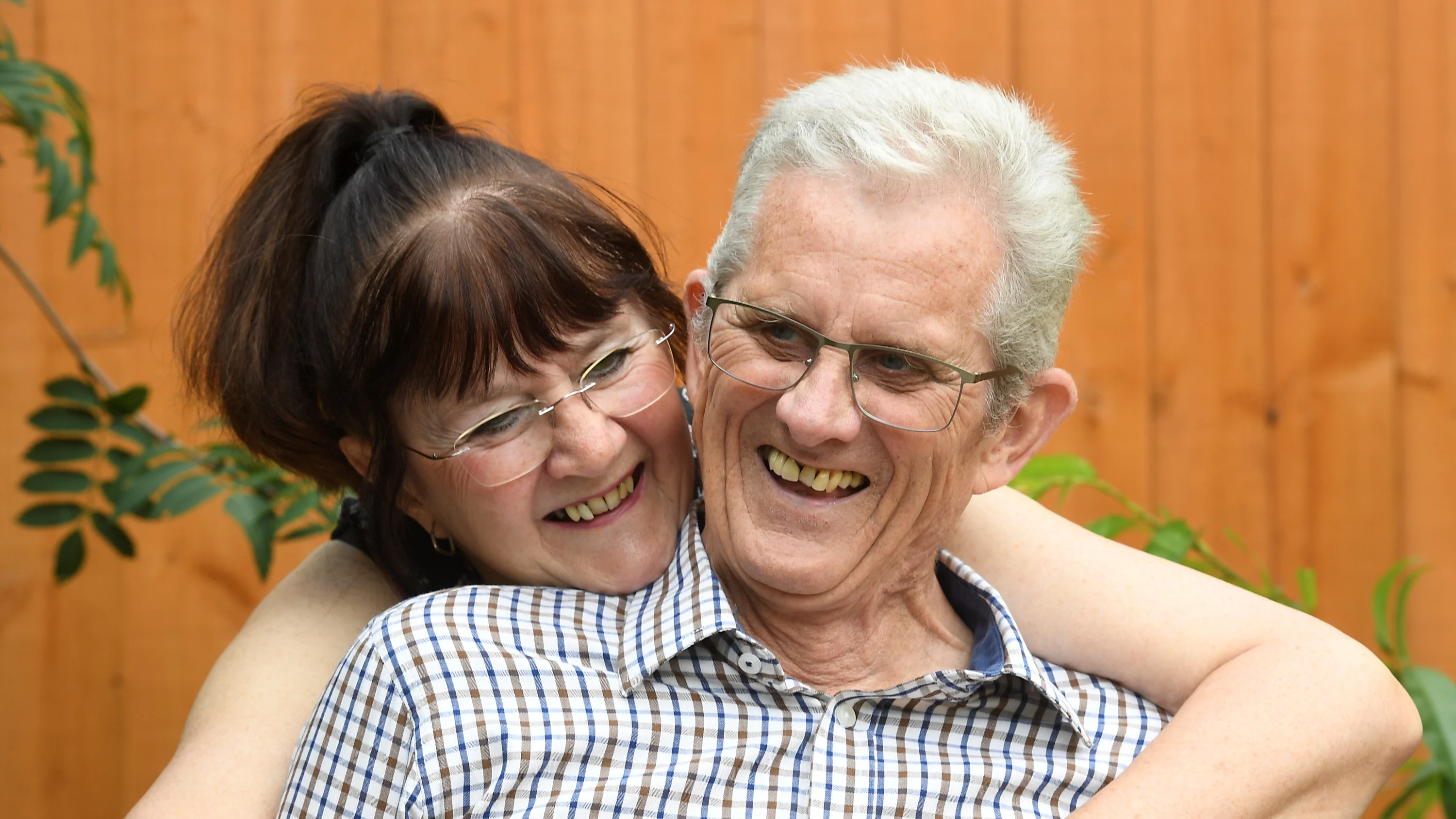 A photograph of Gwen and John in their garden. Gwen is hugging John from behind and they're both laughing.