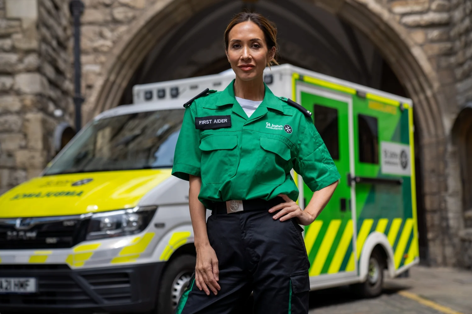 Myleene Klass wearing a St John Ambulance First Aider uniform, stood in front of an ambulance.