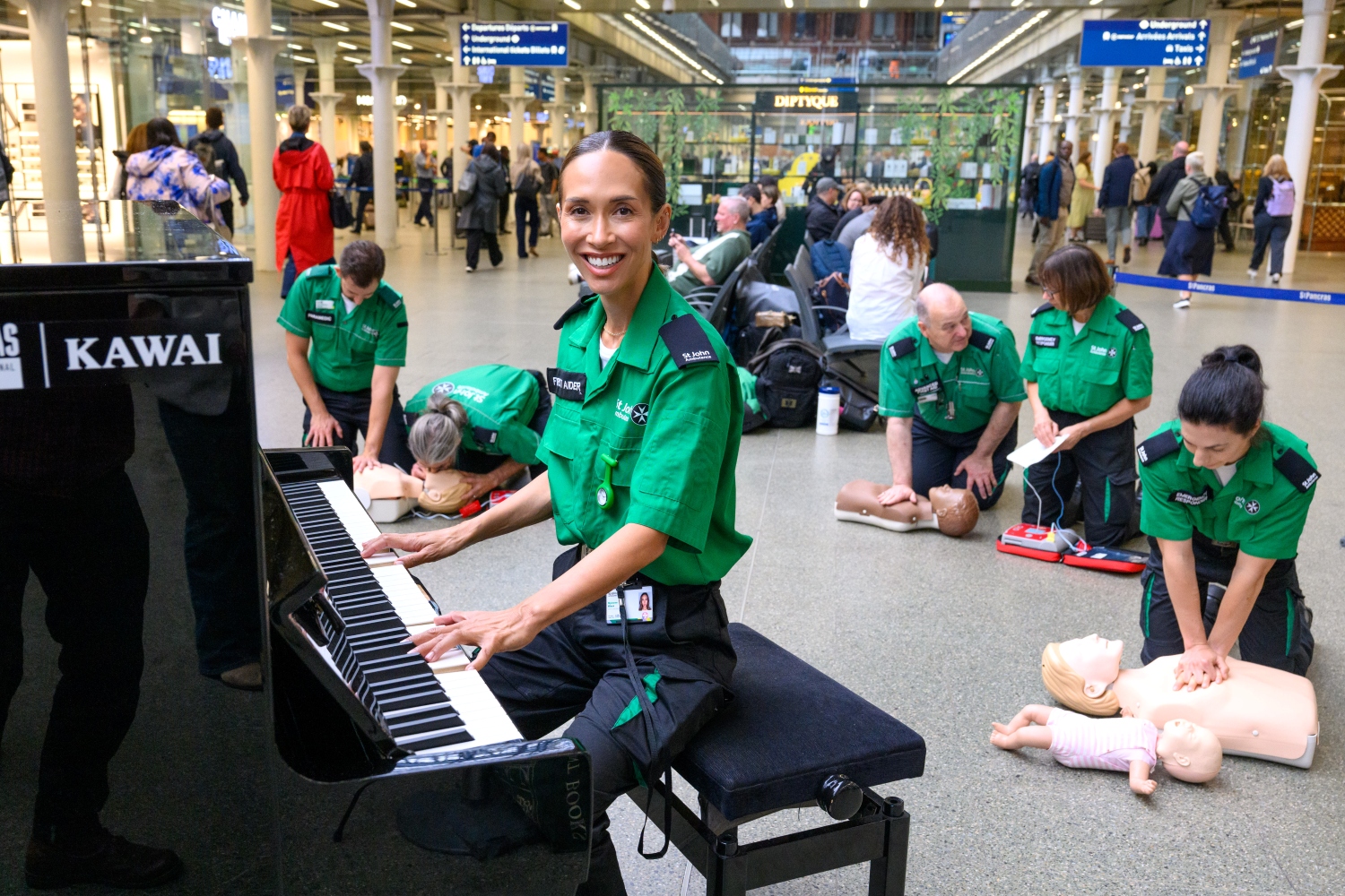 Myleene Klass wearing a First Aider uniform and sat at a piano, smiling. Behind her are five St John Ambulance First Aiders doing chest compressions on CPR mannequins. They are inside St Pancras train station.  