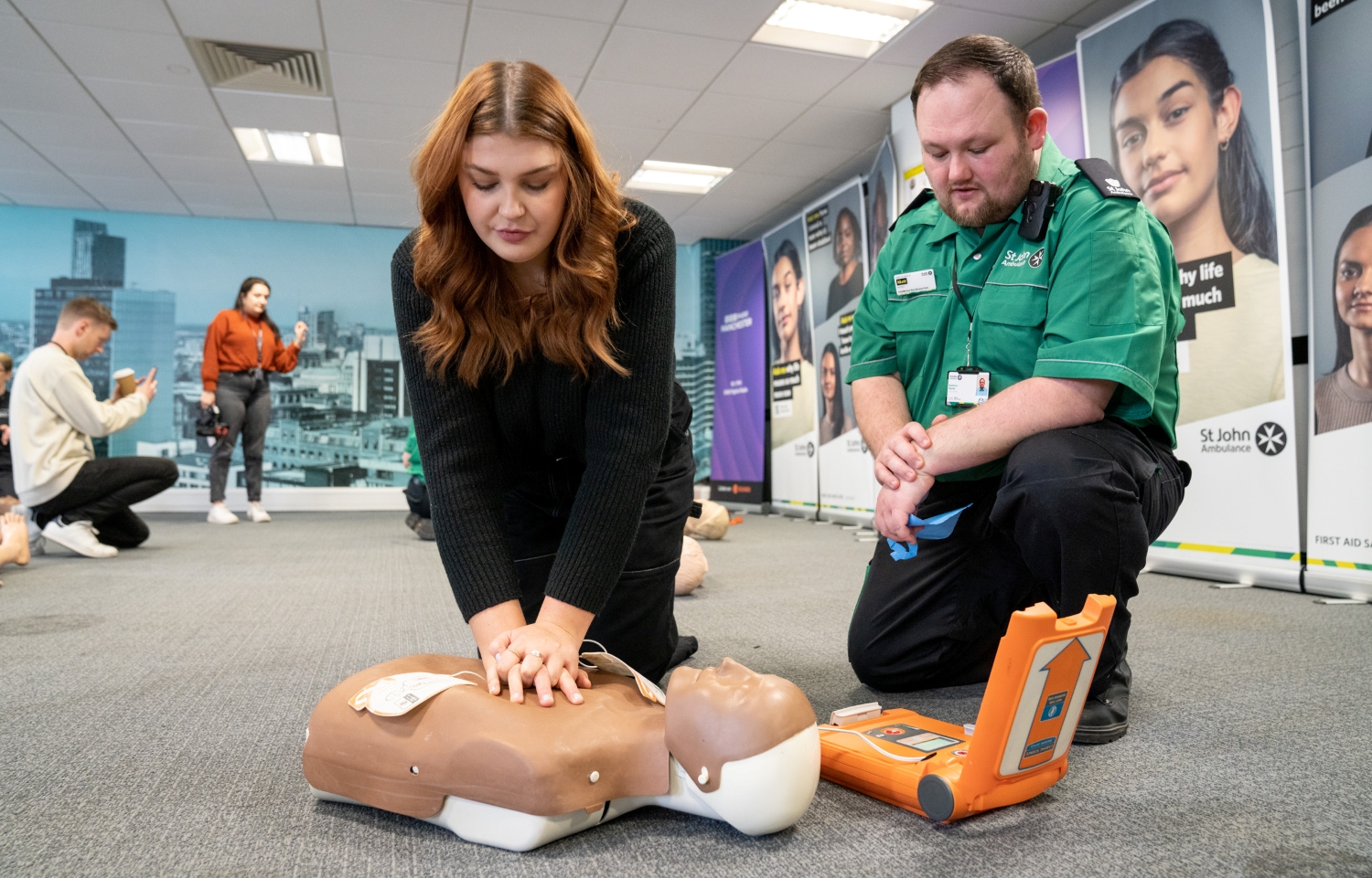 A first aid trainer is kneeling on the floor next to someone doing a chest compression on a mannequin with a defibrillator next to them. They are in a St John Ambulance training room with other people in the background. 