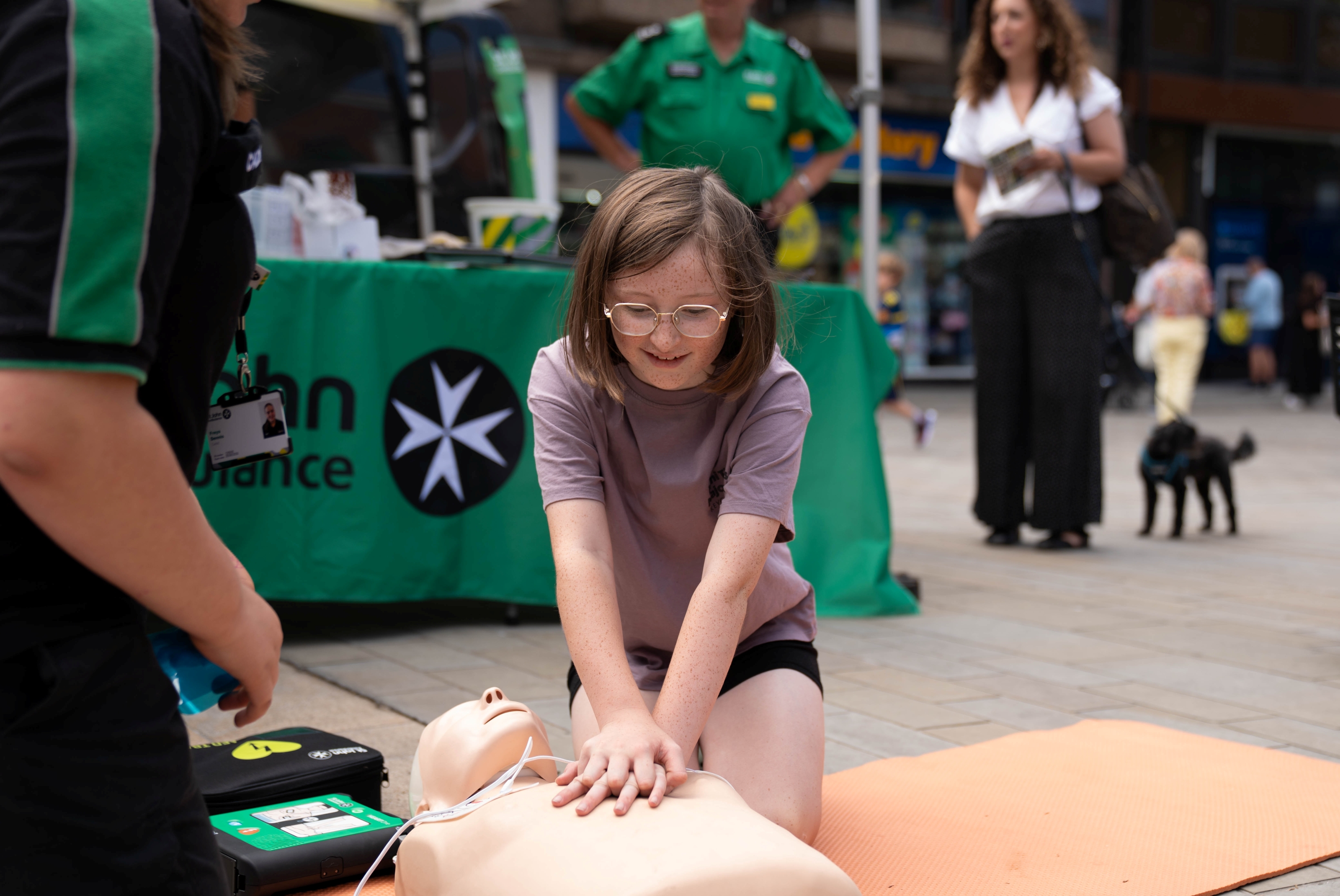 A St John Ambulance volunteer is kneeling on the floor next to a child who is doing chest compressions on a first aid manikin with a defibrillator next to it. Behind them is another volunteer and a member of the public next to a table with 'St John Ambulance' on it. 
