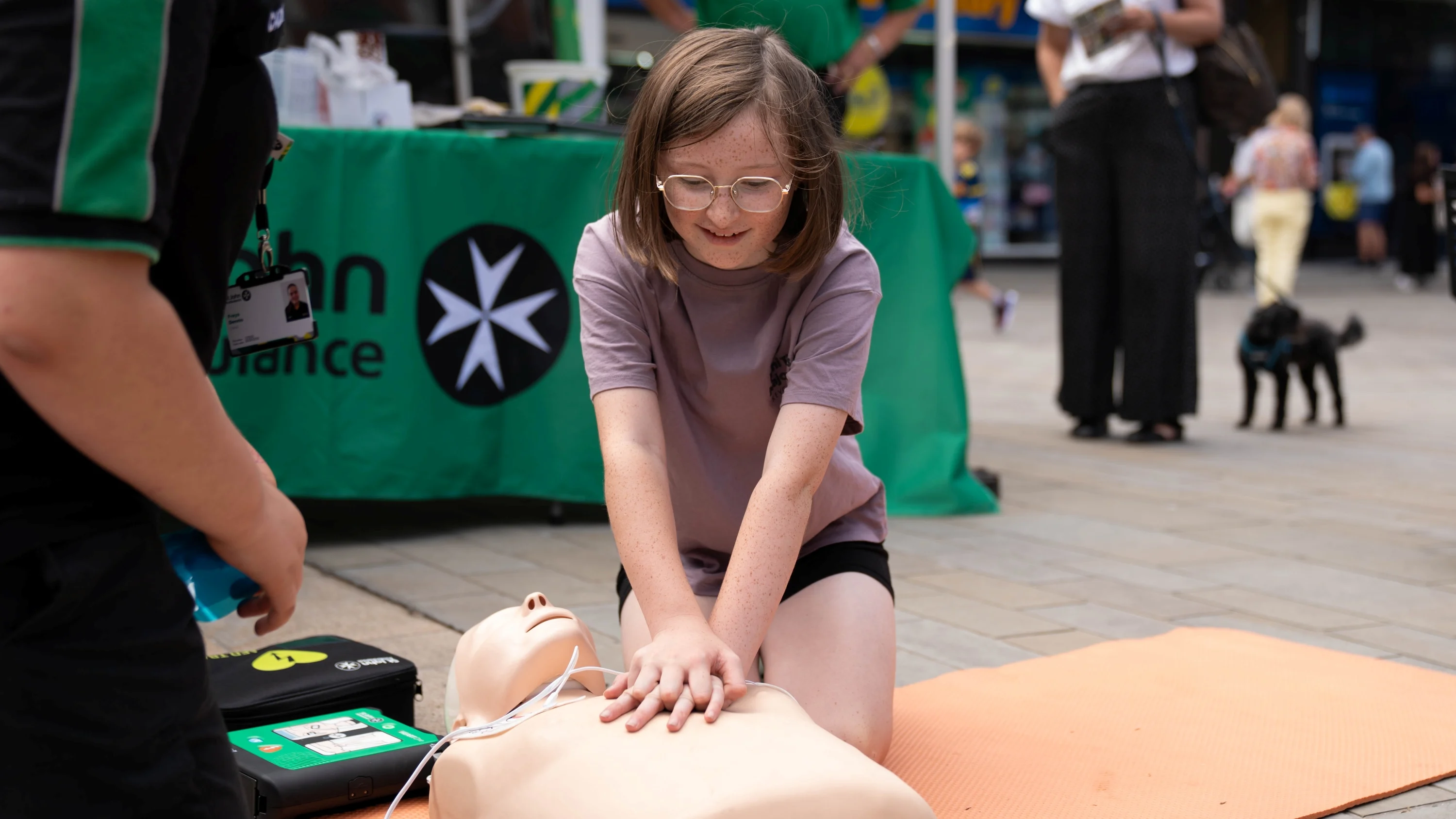 A St John Ambulance volunteer is kneeling on the floor next to a child who is doing chest compressions on a first aid manikin with a defibrillator next to it. Behind them is another volunteer and a member of the public next to a table with 'St John Ambulance' on it.
