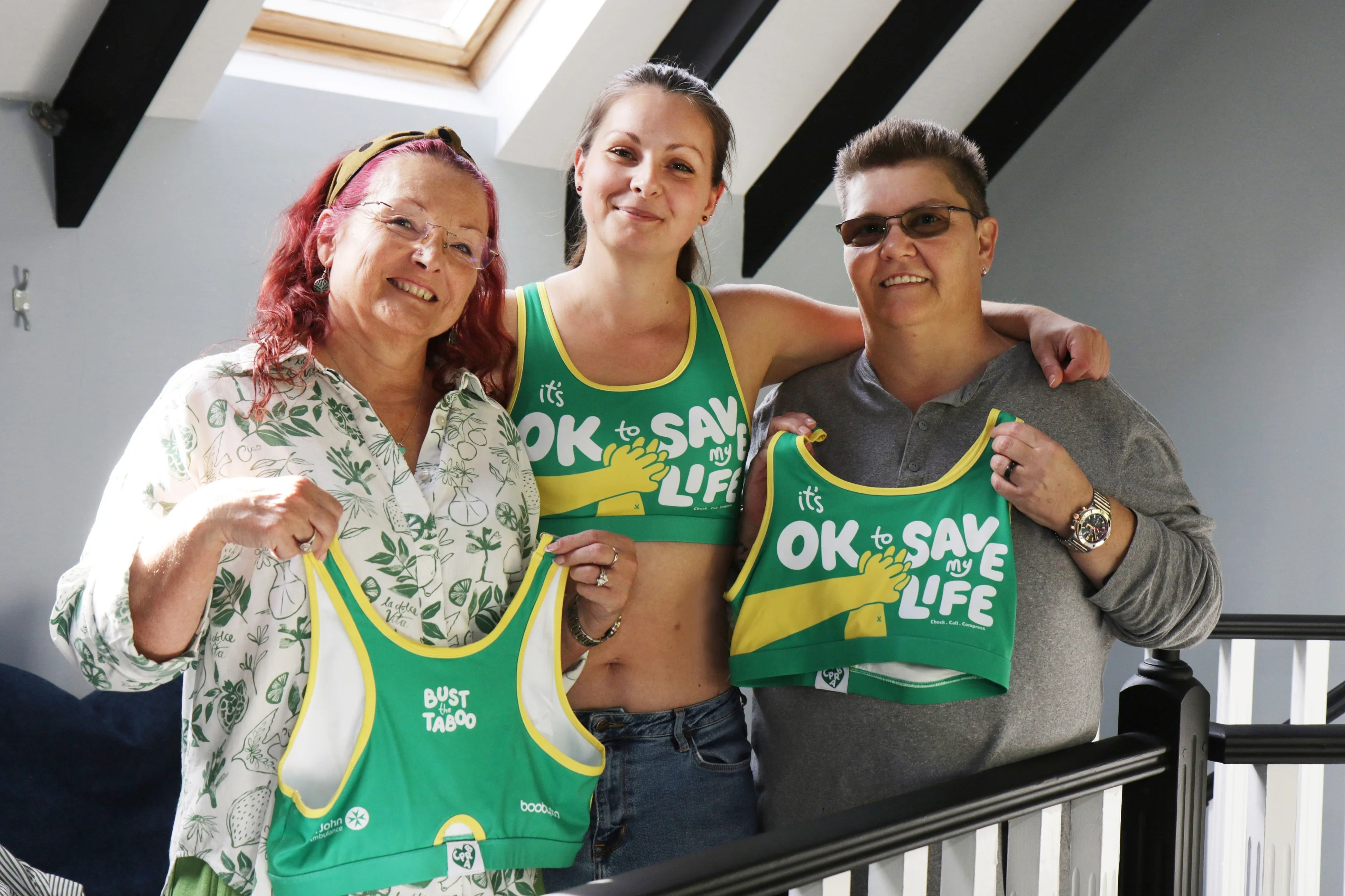 Sally and Carole holding up a CPR Bra with Jade, Sally's daughter, stood in the middle of them wearing a CPR Bra. On the front of the bra, it says 'It's ok to save my life'. 