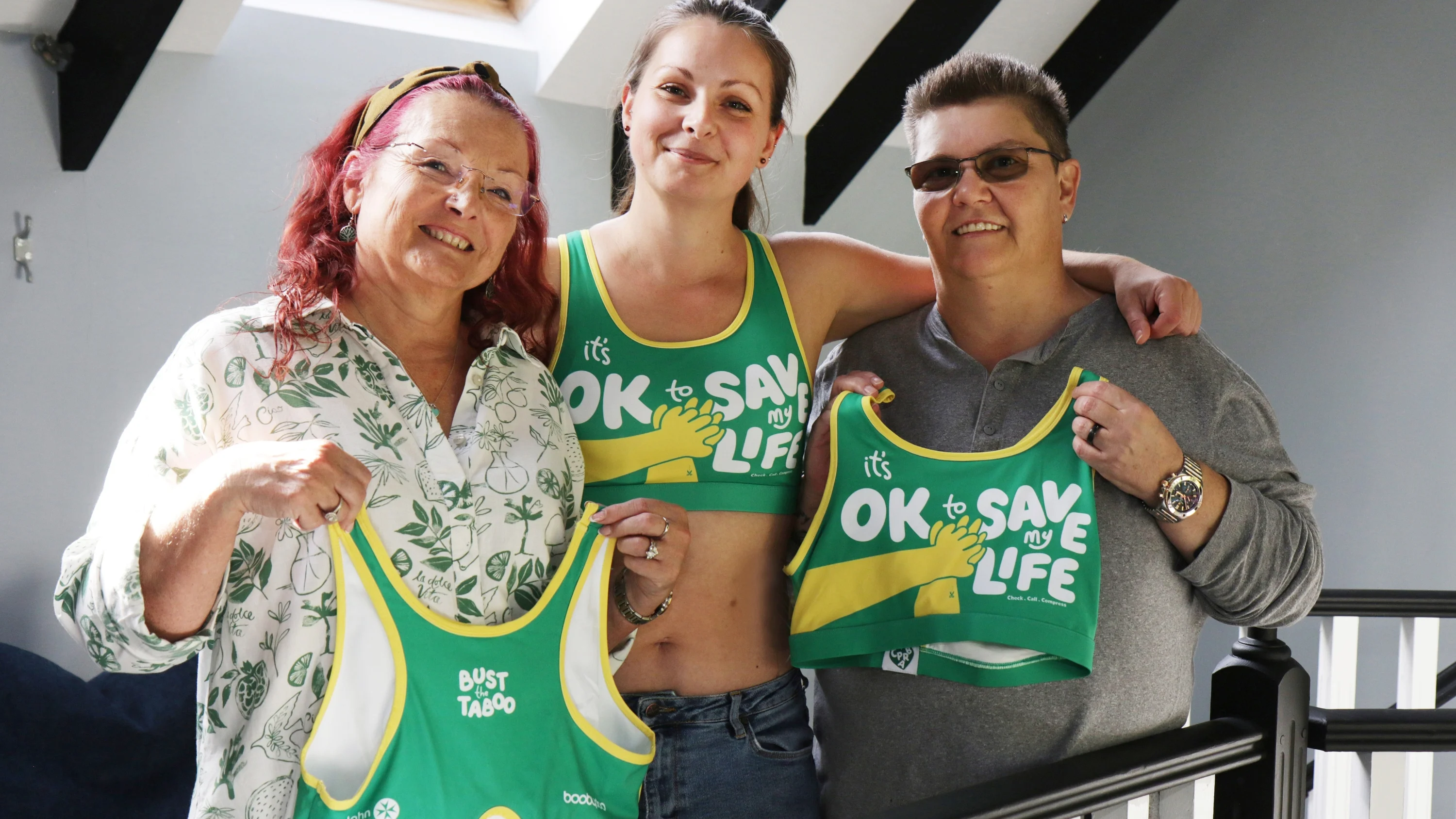Sally and Carole holding up a CPR Bra with Jade, Sally's daughter, stood in the middle of them wearing a CPR Bra. On the front of the bra, it says 'It's ok to save my life'.