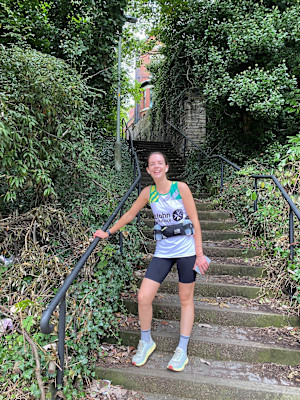 A photograph of Alice in her running gear and St John vest, posing for the camera on stone steps, surrounded by greenery.