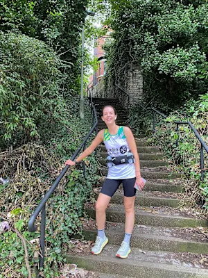 A photograph of Alice in her running gear and St John vest, posing for the camera on stone steps, surrounded by greenery.