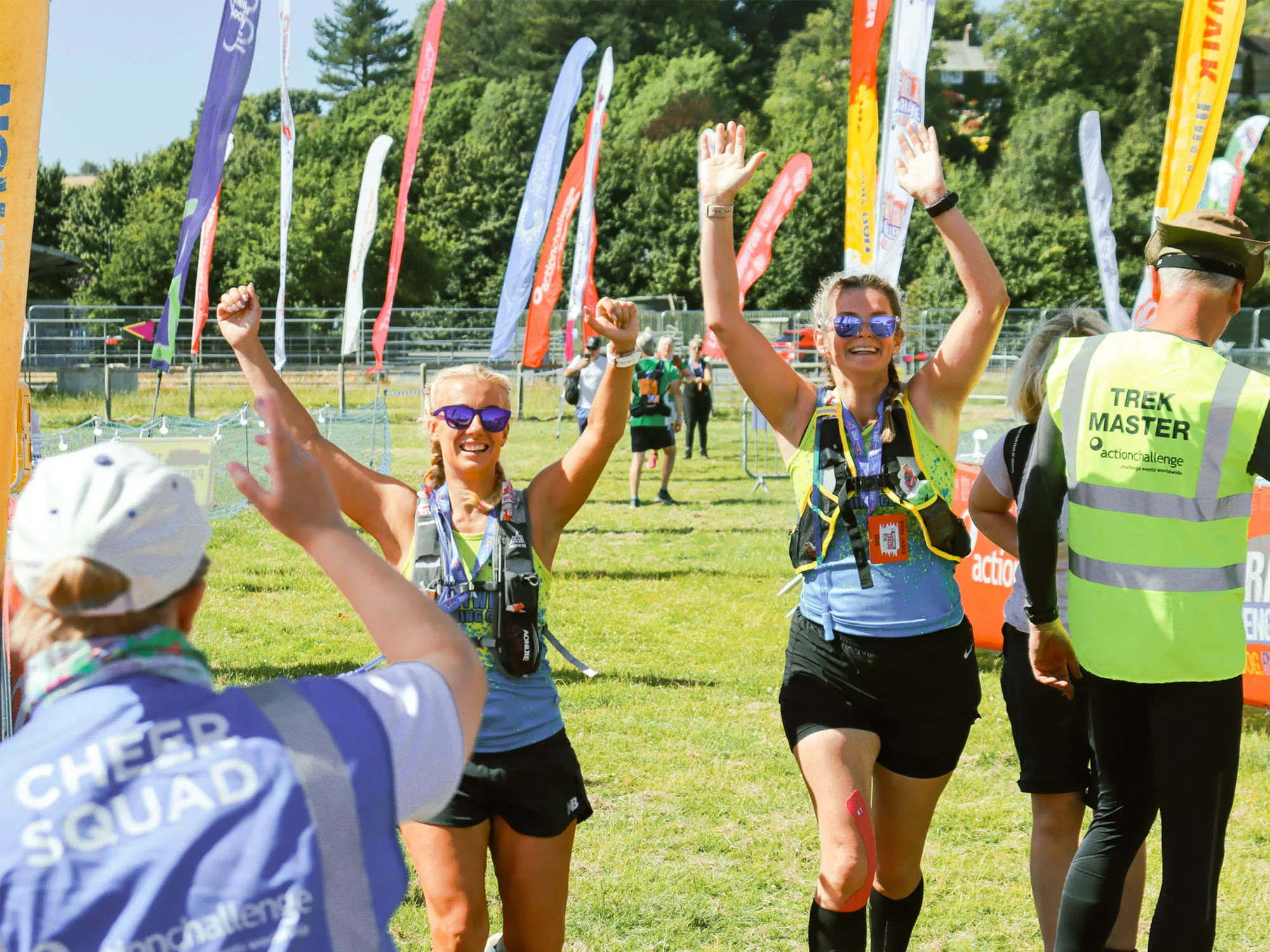 Two people with their arms in the air walking across a field with lots of tall flags and other people standing around. Another person is in front of them wearing a blue hi-vis vest with 'Cheer squad' on the back of it.