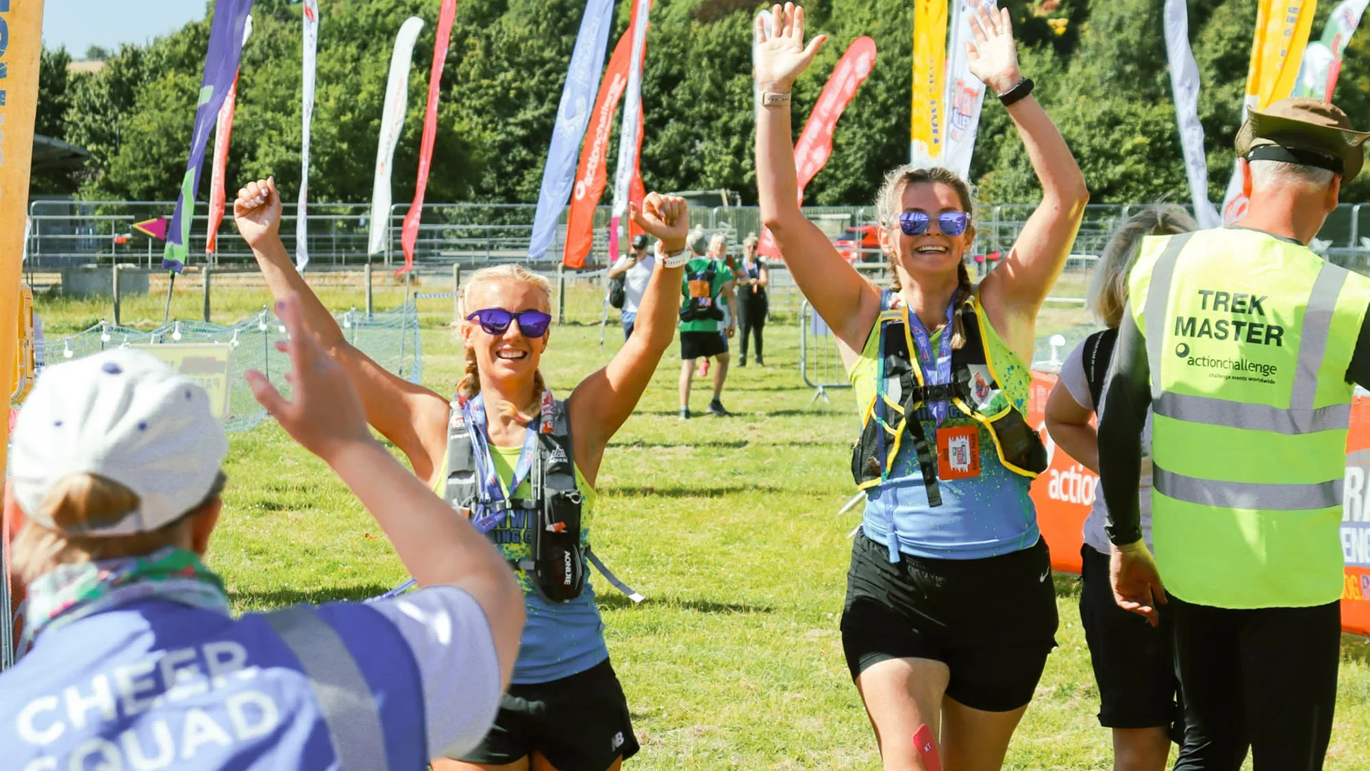 Two people with their arms in the air walking across a field with lots of tall flags and other people standing around. Another person is in front of them wearing a blue hi-vis vest with 'Cheer squad' on the back of it.