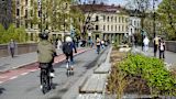 People riding bicycles along a paved city path lined with buildings and trees.