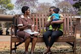 Two women talking on an outdoor bench, one holding a baby