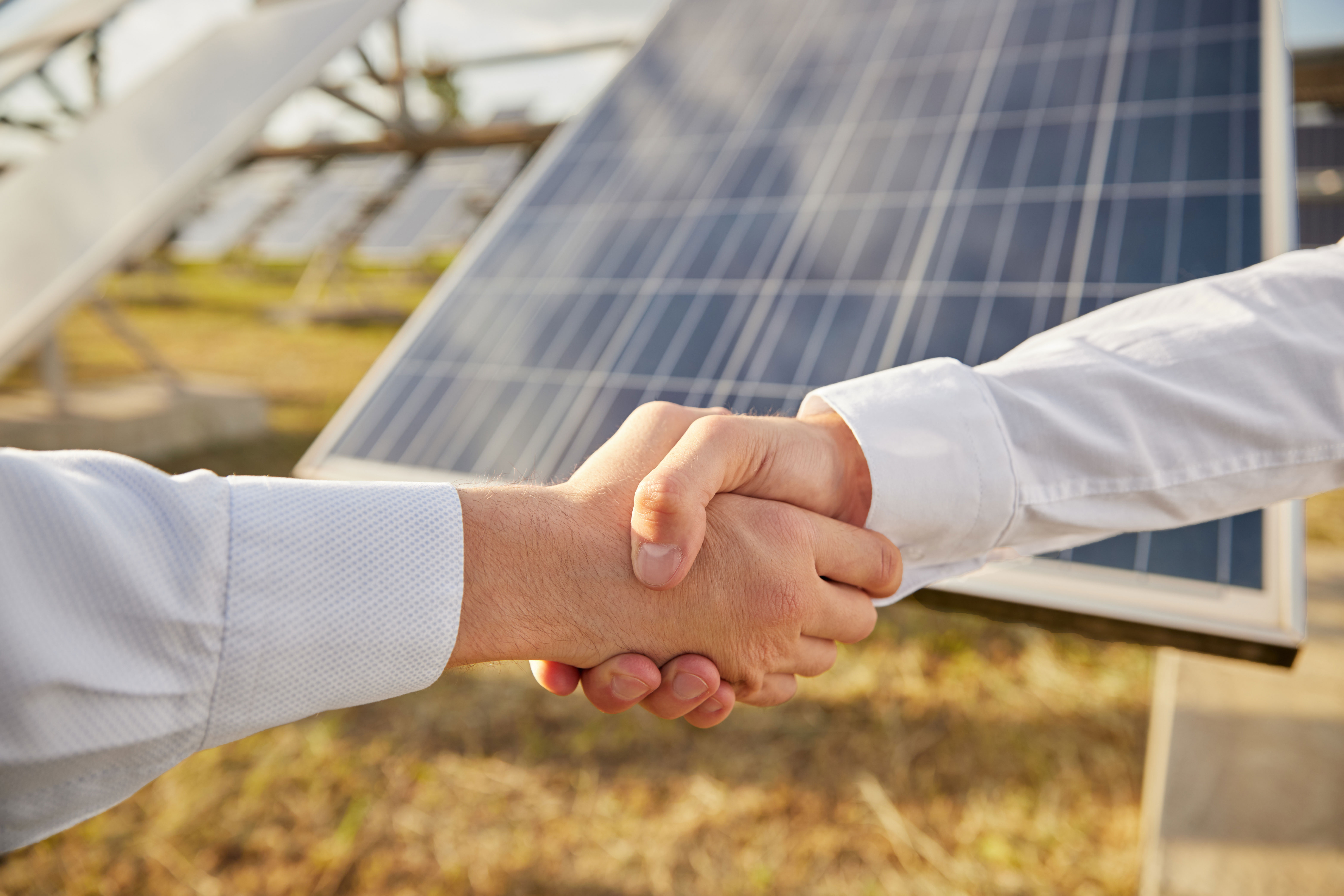 Zwei Geschäftspartner geben sich die Hand. Im Hintergrund ist ein Solarpark angedeutet.