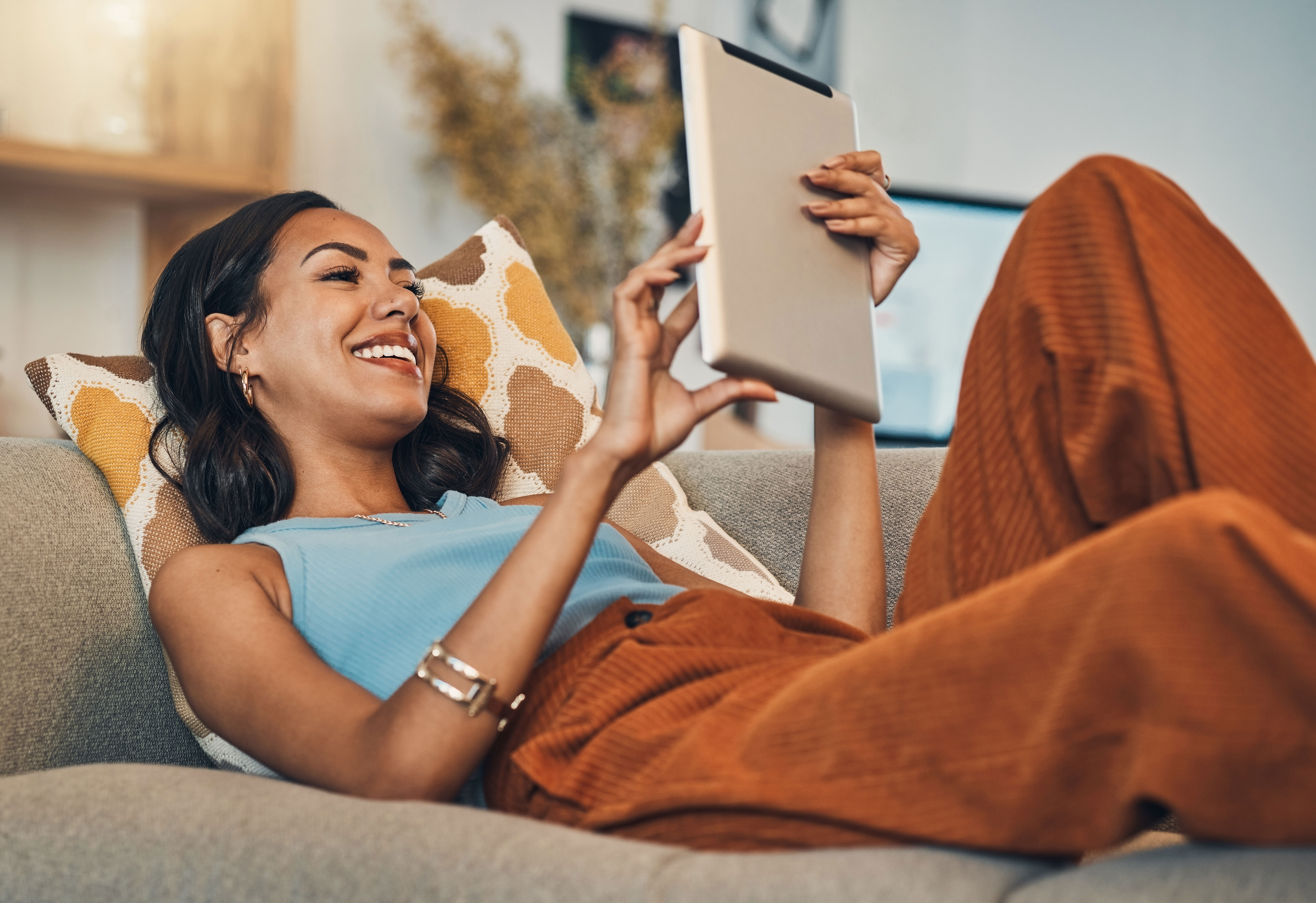Frau liegt auf einem Sofa und hält lächelnd ein Tablet in der Hand.