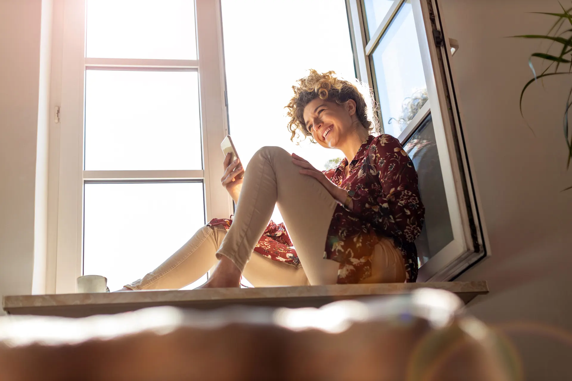 Attraktive Tarife bei der NEW Energie Frau mit einem Smartphone in der Hand sitzt auf einer Fensterbank vor dem geöffneten Fenster.
