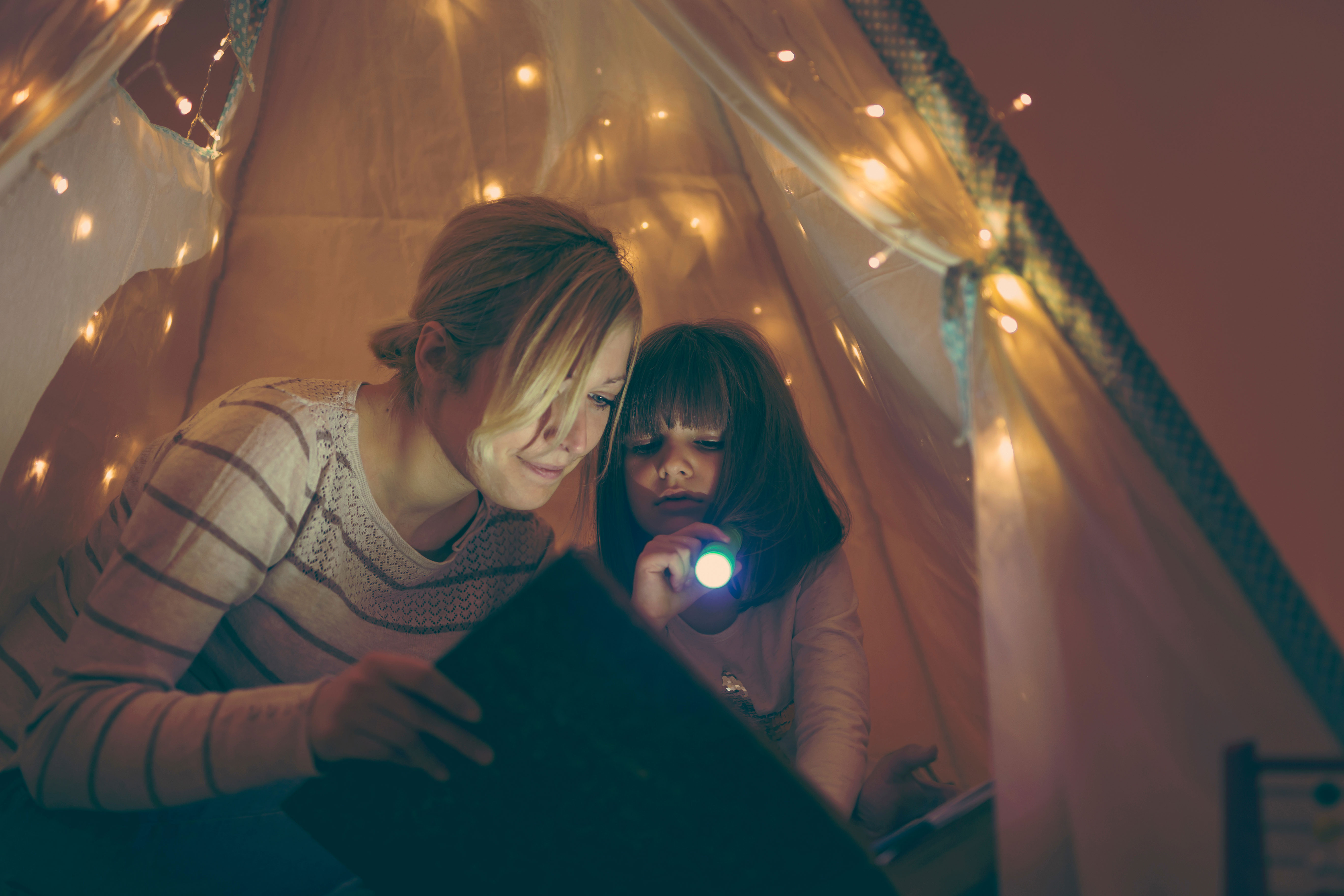 Mutter sitzt mit ihrer Tochter in einem beleuchteten Zelt und hat eine Taschenlampe in der Hand. Gemeinsam lesen sie ein Buch.