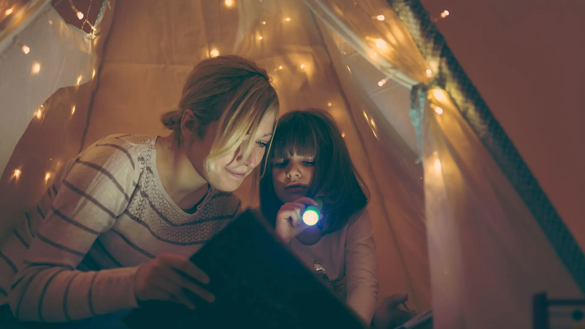 Was Sie bei einem Stromausfall tun können Mutter sitzt mit ihrer Tochter in einem beleuchteten Zelt und hat eine Taschenlampe in der Hand. Gemeinsam lesen sie ein Buch.
