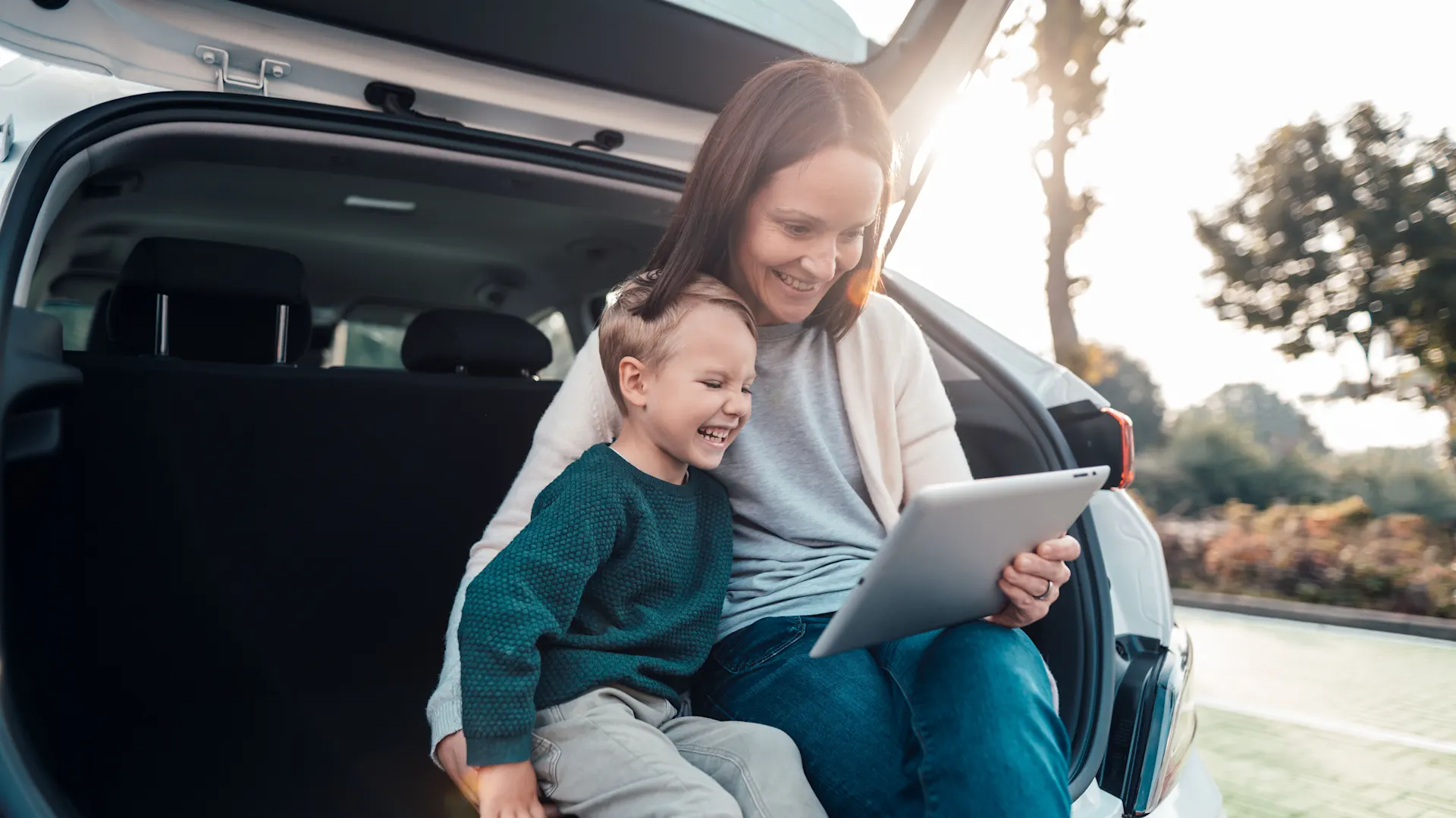 Familie informiert sich über Produkte Eine Mutter mit Kind parkt auf einem öffentlichen Parkplatz, sitzt im Kofferraum und informiert sich mit einem Tablet über den Energieversorger NEW Energie.