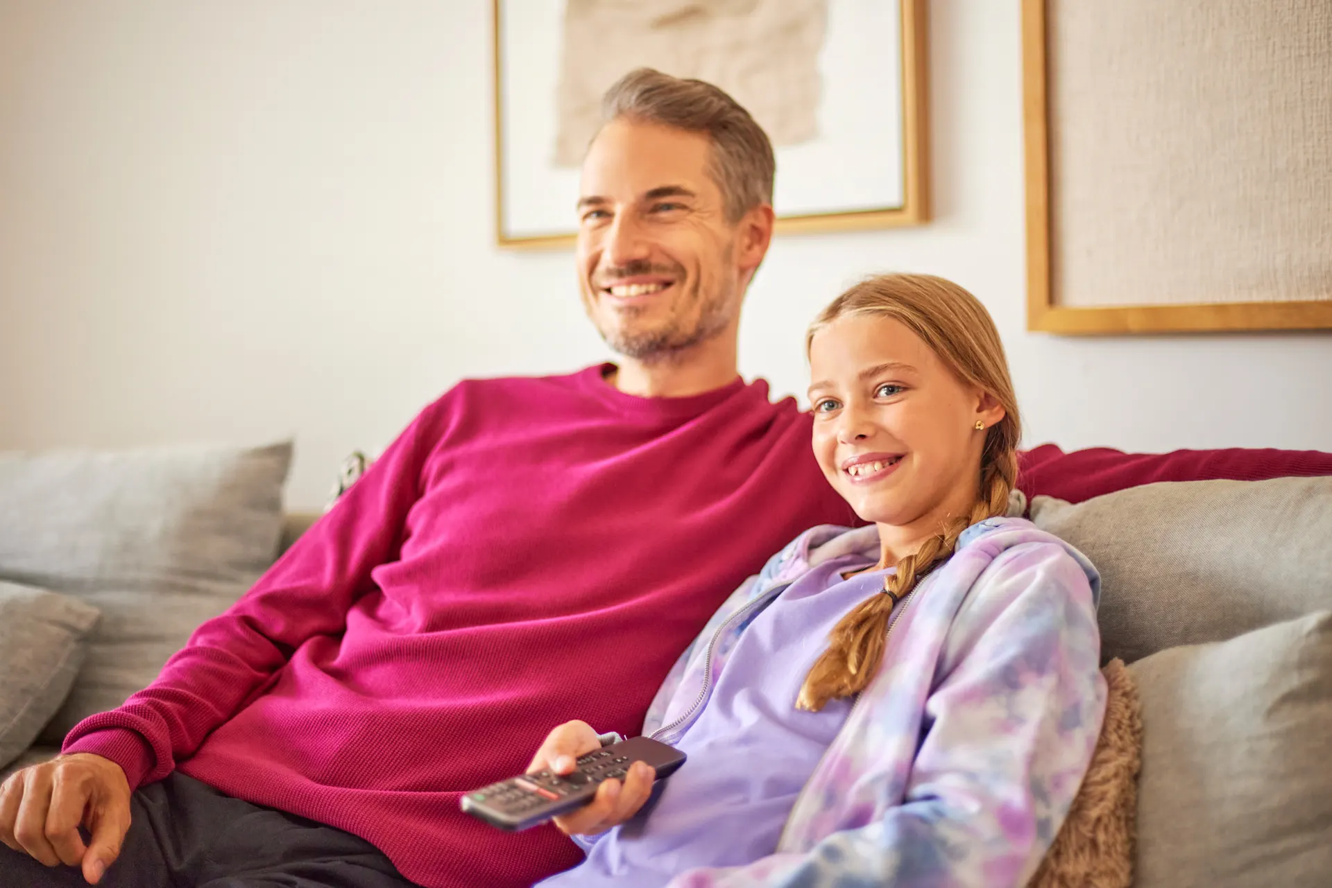 gemeinsam mit der Familie Zeit verbringen Ein Mann sitzt mit seiner Tochter auf dem Sofa im Wohnzimmer. Sie schauen etwas im Fernsehen. Dieser wird mit Strom von der NEW Energie betrieben.