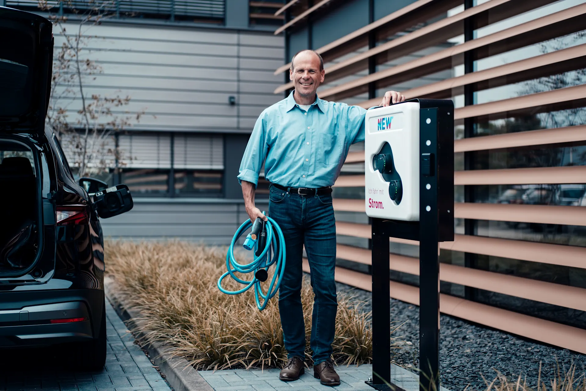 Mann angelehnt an einer Wallbox Ein Mann steht lächelnd neben einem Elektroauto an einer Wallbox und hält ein Ladekabel in der Hand.