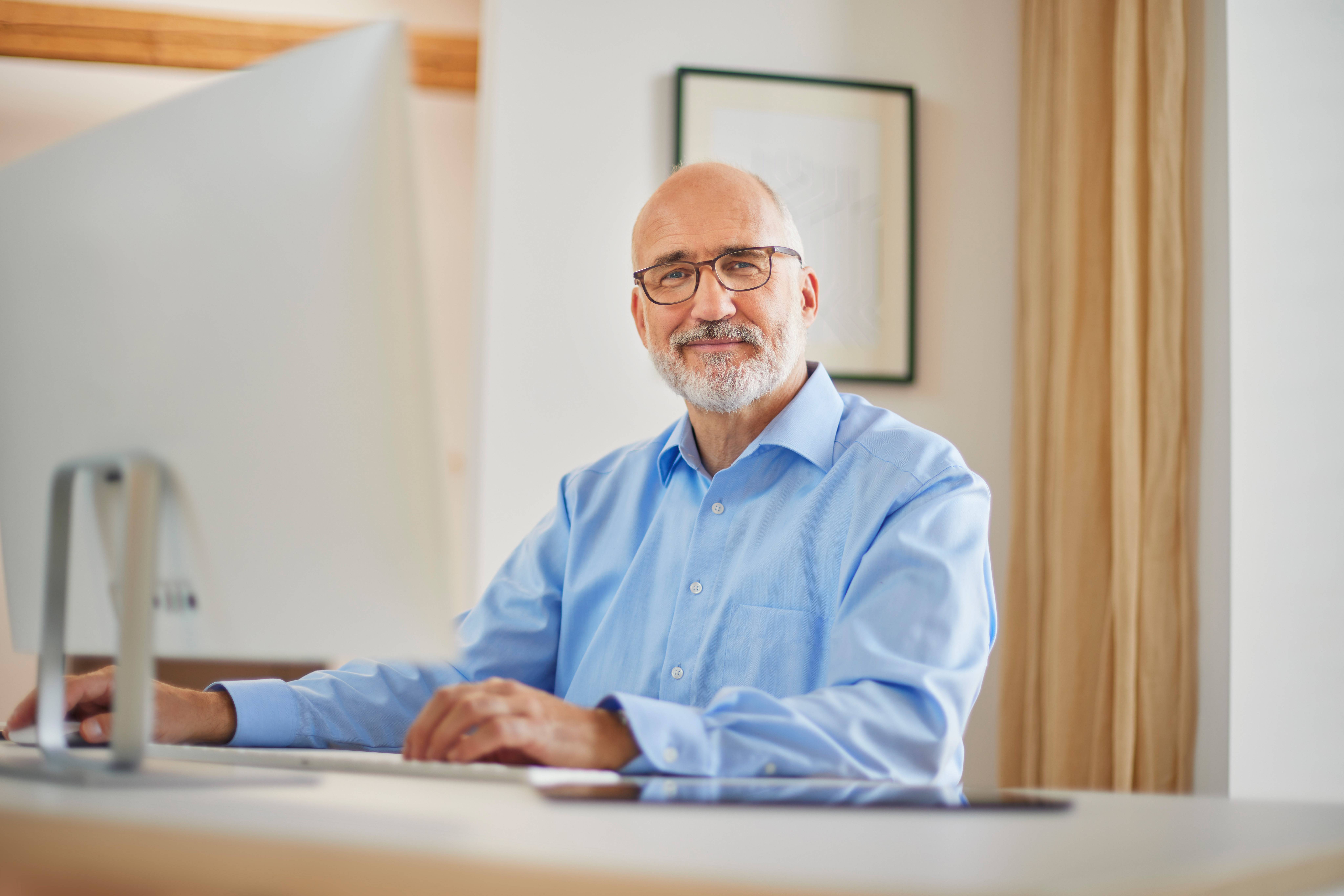 Ein Hausverwalter sitzt am Schreibtisch und arbeitet am Computer und lächelt dabei leicht.