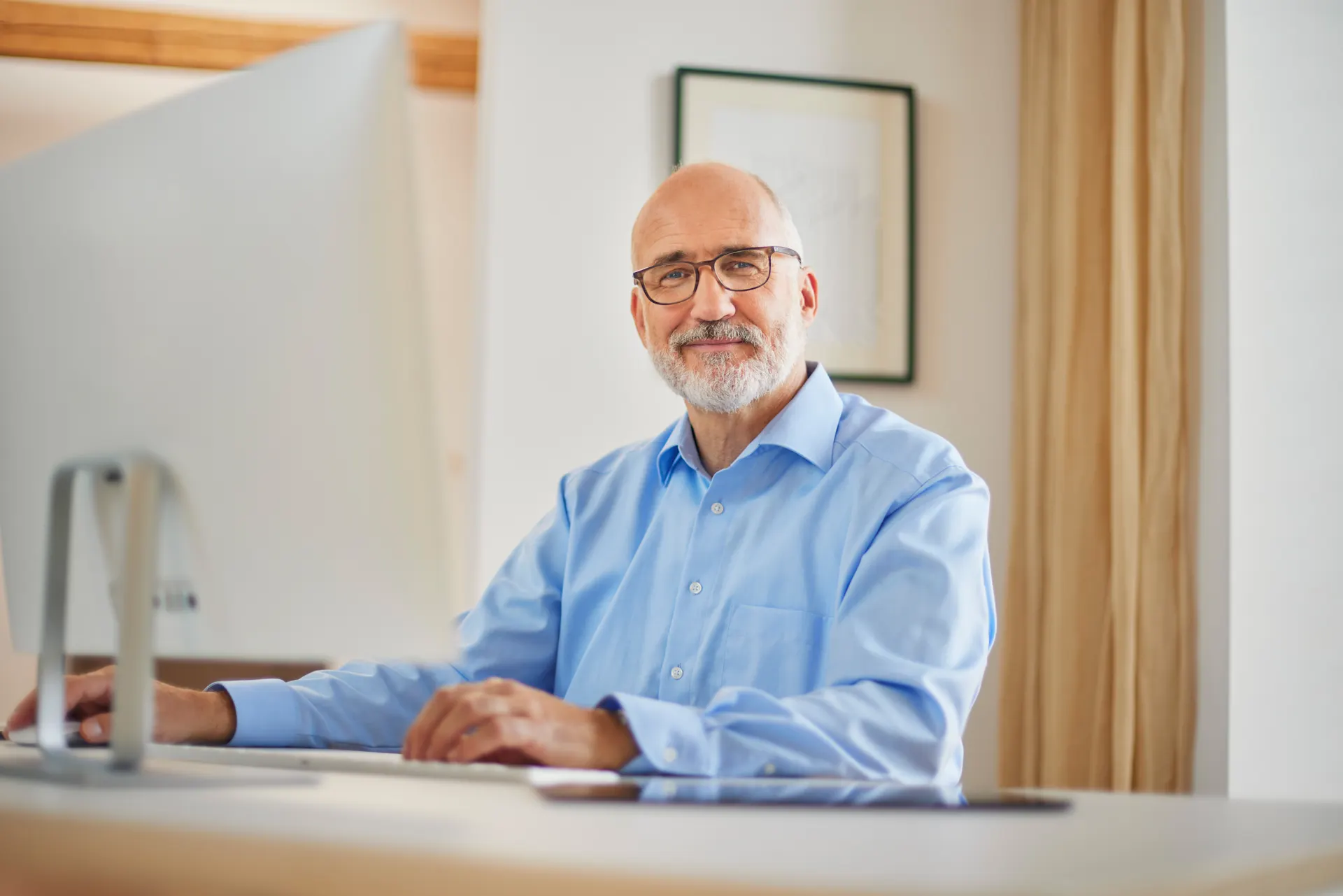 Ein Hausverwalter sitzt am Schreibtisch und arbeitet am Computer und lächelt dabei leicht.
