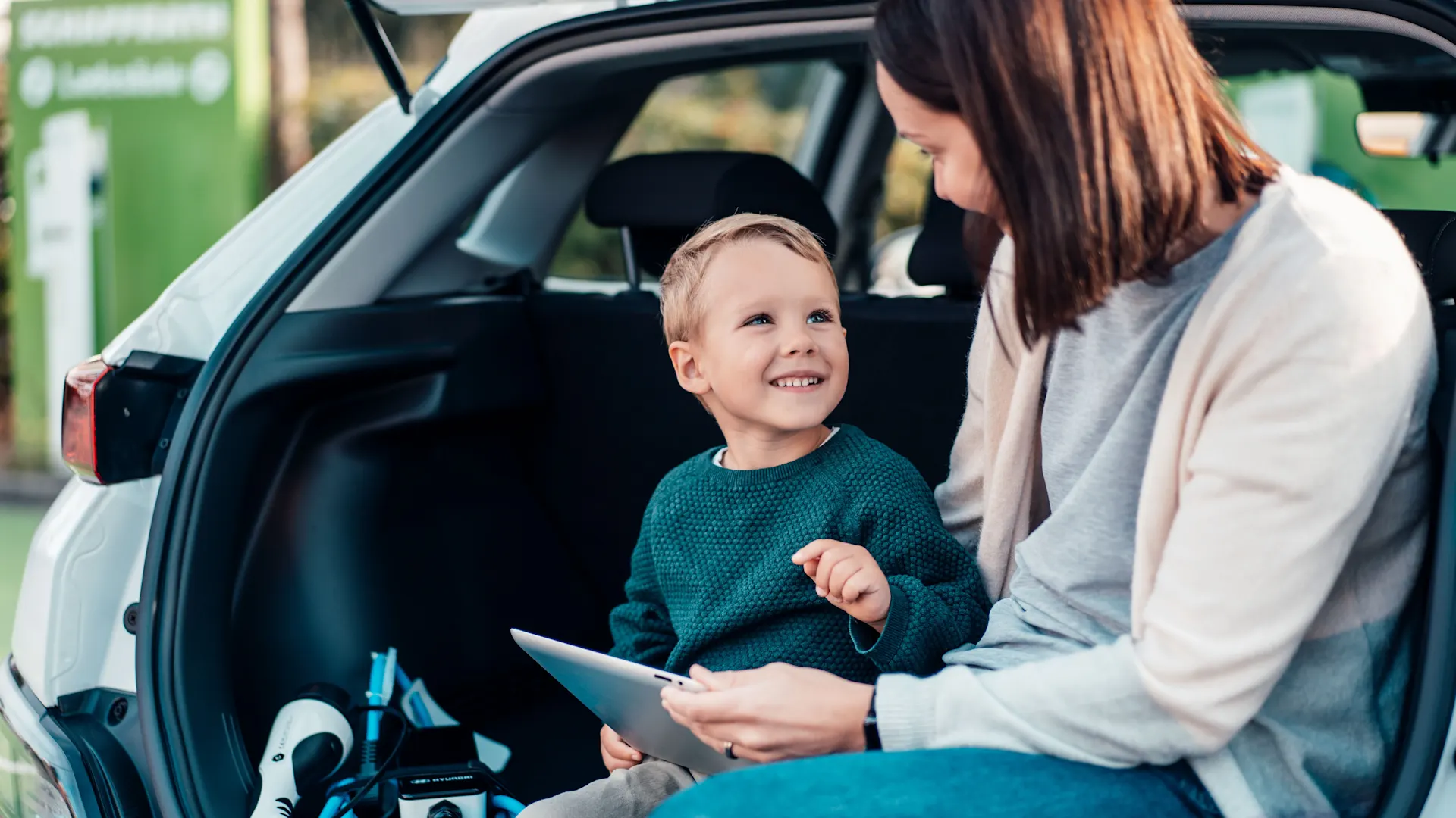 Familie informiert sich über Wissenswertes der NEW Energie Mutter und Sohn lächeln sich im offenen Kofferraum an und schauen übers Tablet nach Informationen über die NEW Energie.