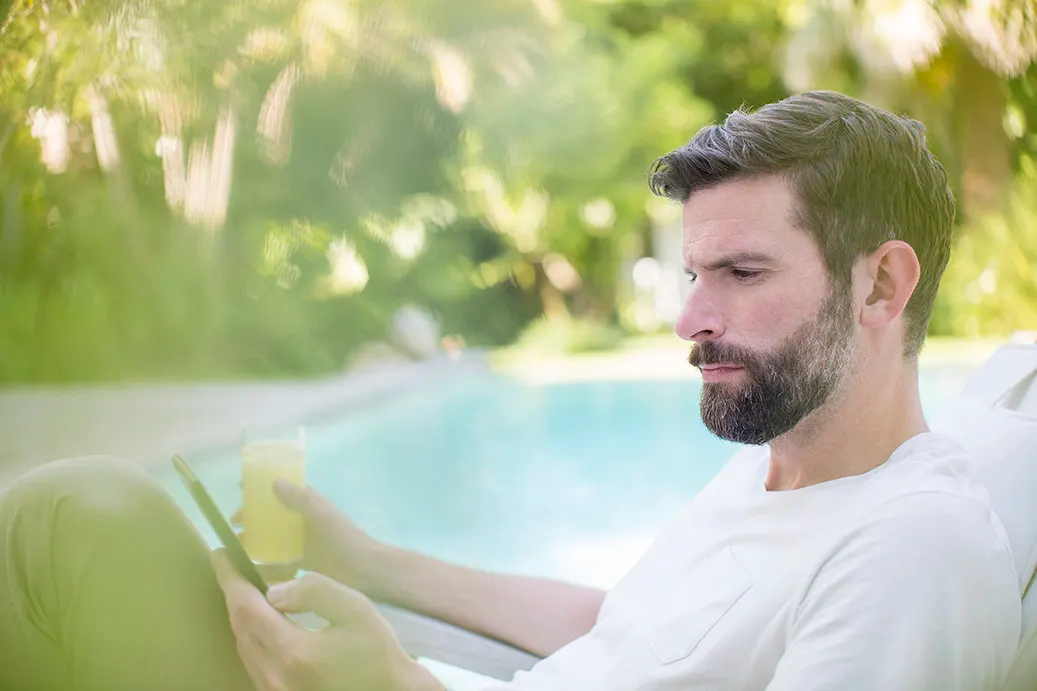 Ein Mann sitzt an einem Pool mit seinem Handy und einem Getränk in der Hand. Dabei schaut er nach Stromtarifen der NEW Energie. 