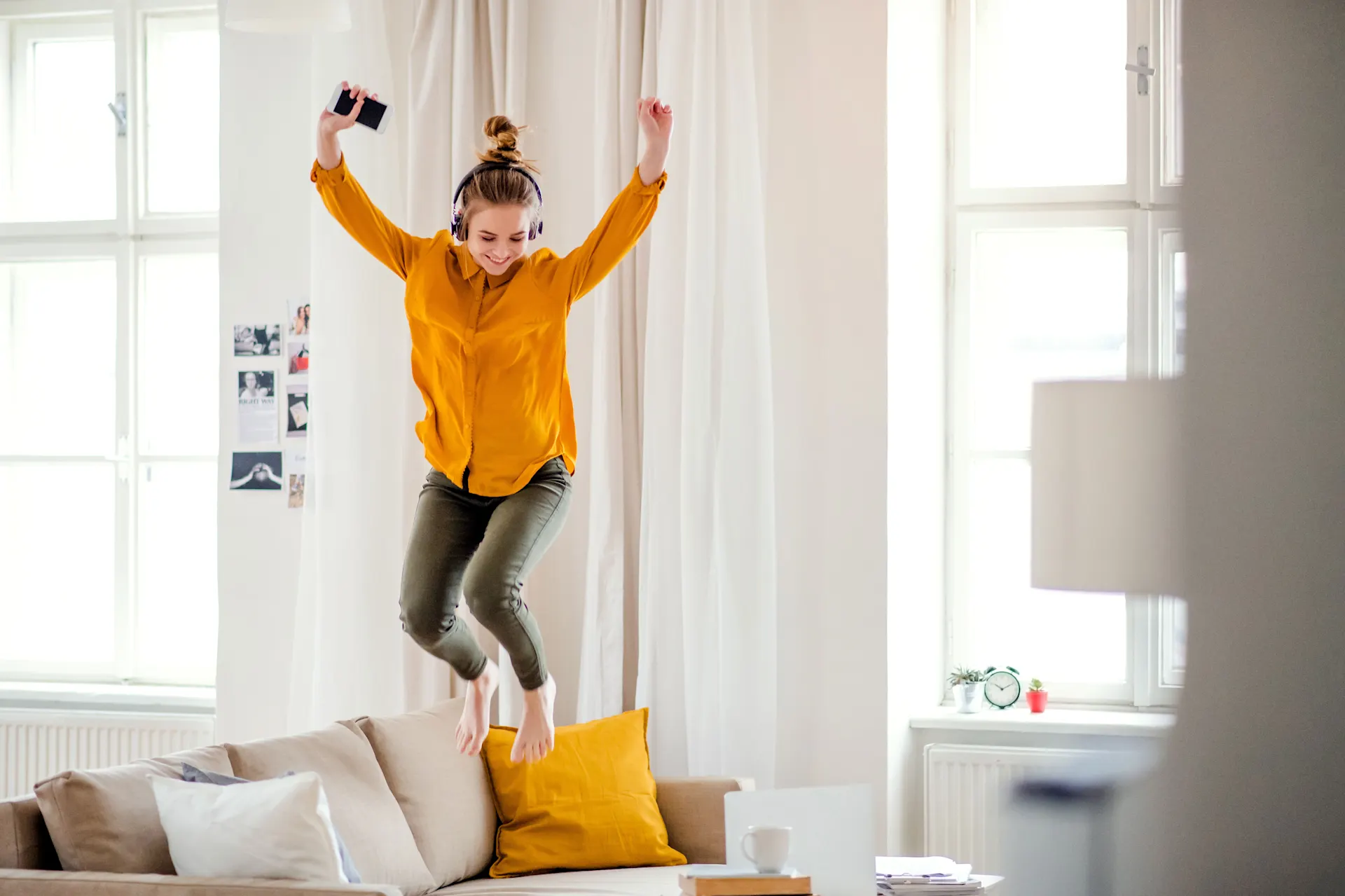 Eine Frau springt glücklich mit einem Smartphone in der Hand und Kopfhörern auf dem Kopf auf dem Sofa rum und freut sich über die NEW Energie.
