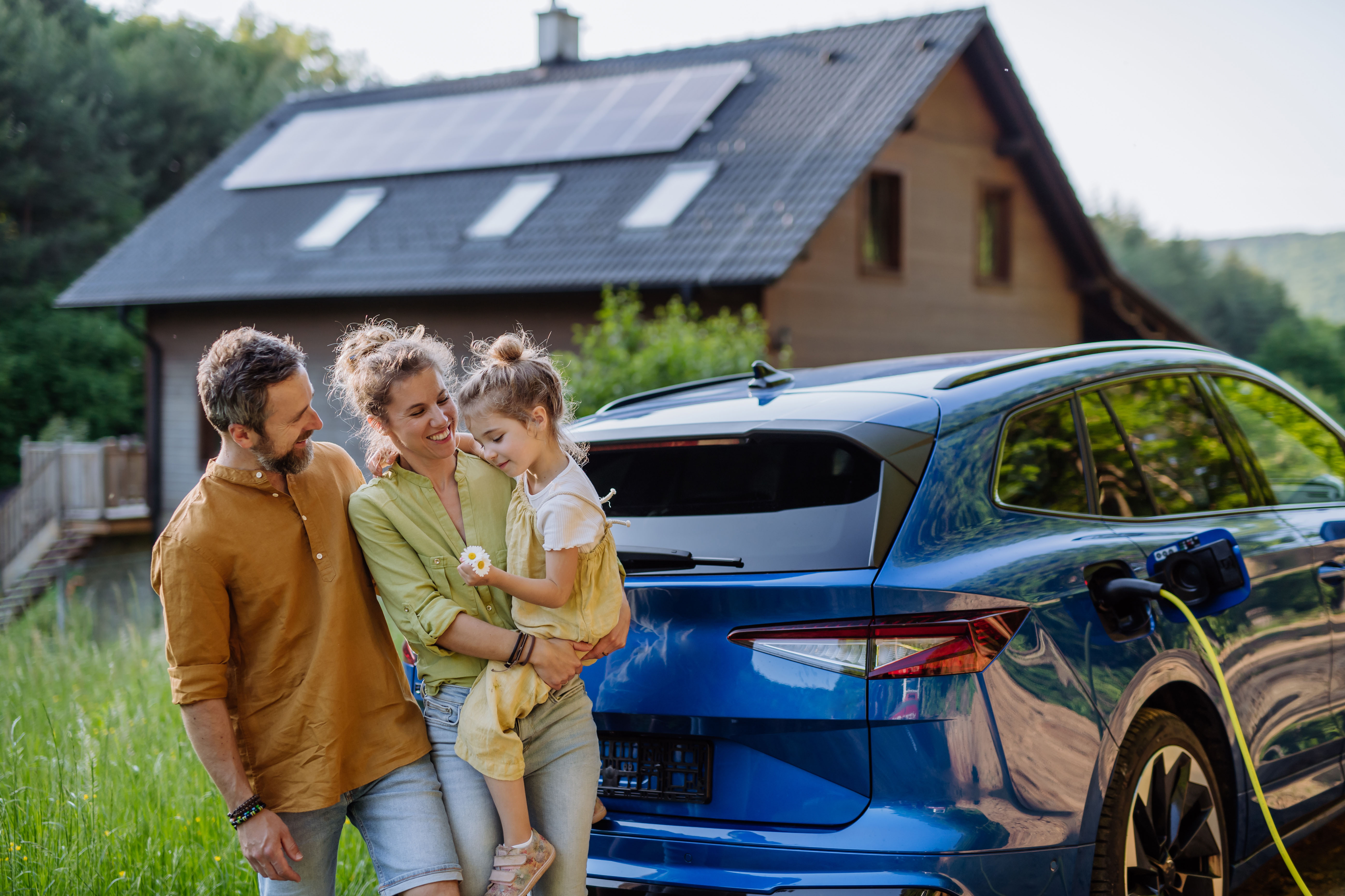 Eine Familie steht vor ihrem E-Auto. Im Hintergrund steht ihr Haus mit einer PV-Anlage der NEW Energie. Ebenfalls beziehen Sie Strom und Gas von der NEW Energie. 