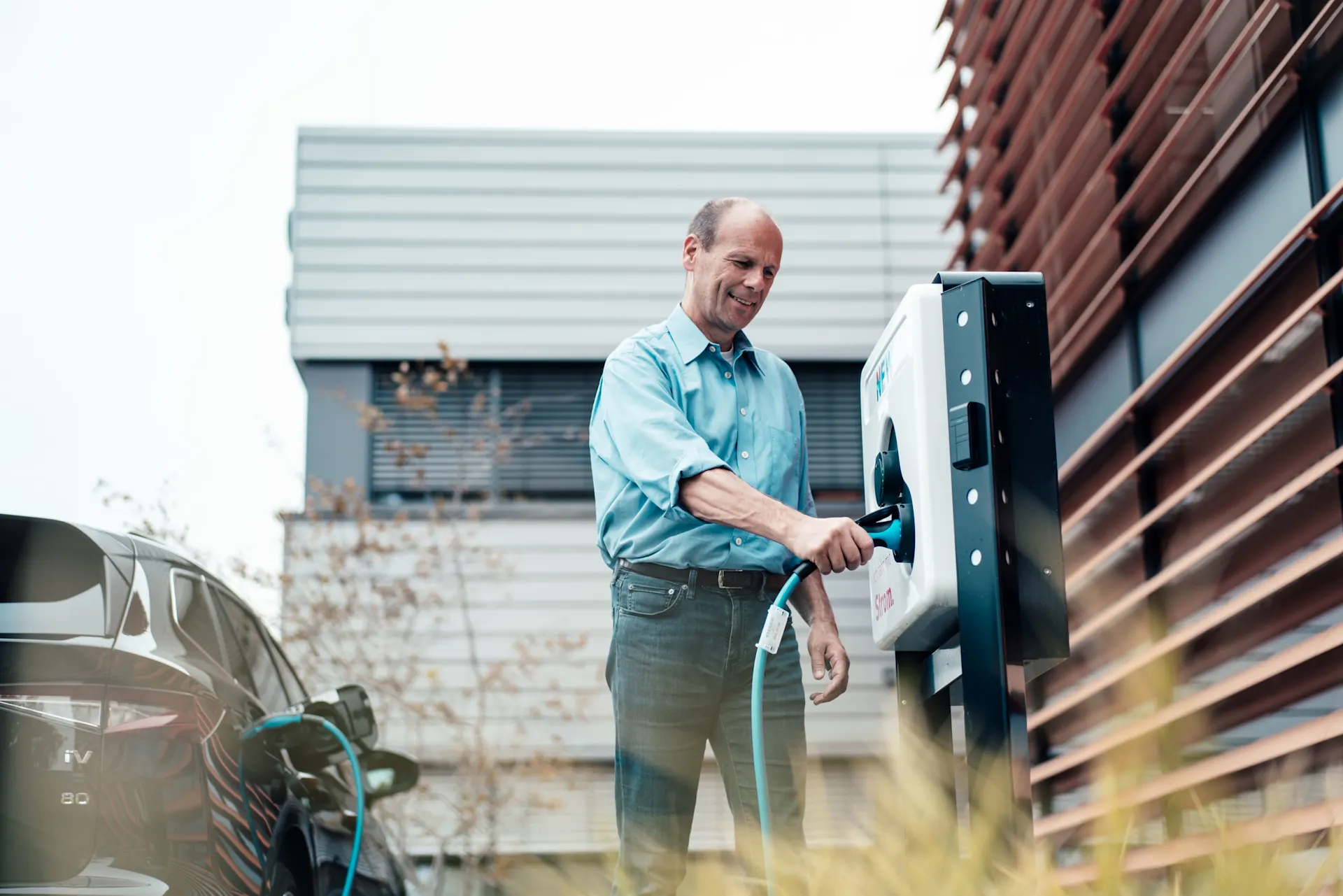 Mann steckt das Ladekabel in eine Wallbox Ein Mann steckt ein Ladekabel in eine Wallbox. Die Wallbox steht an einem Geschäftsgebäude.