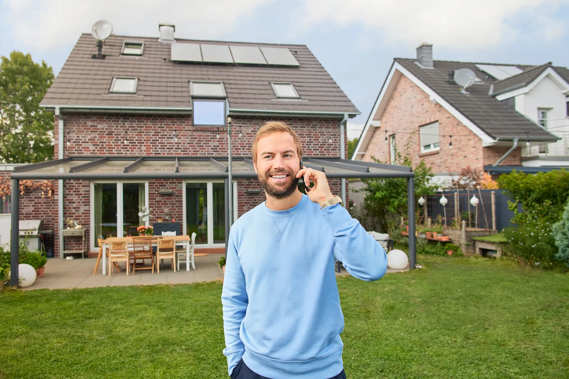 Photovoltaikanlage kaufen Ein Mann telefoniert mit seinem Handy im Garten vor seinem Einfamilienhaus, auf dessen Dach eine Photovoltaikanlage der NEW-Energie angebracht ist.