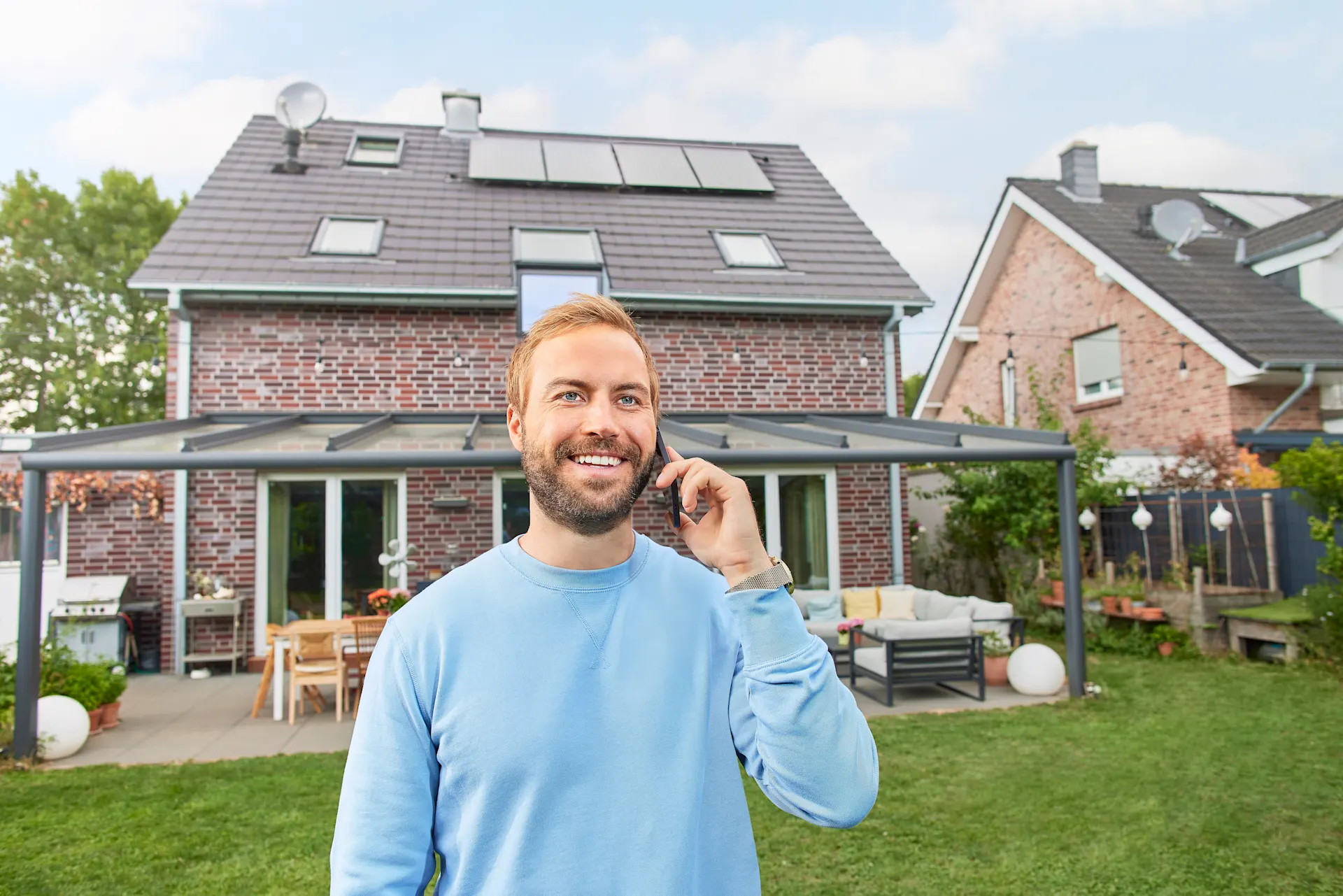 Ein zufriedener Photovoltaikanlagen-Besitzer telefoniert vor seinem Einfamilienhaus, auf dessen Dach die Photovoltaikanlage der NEW Energie zu sehen ist.