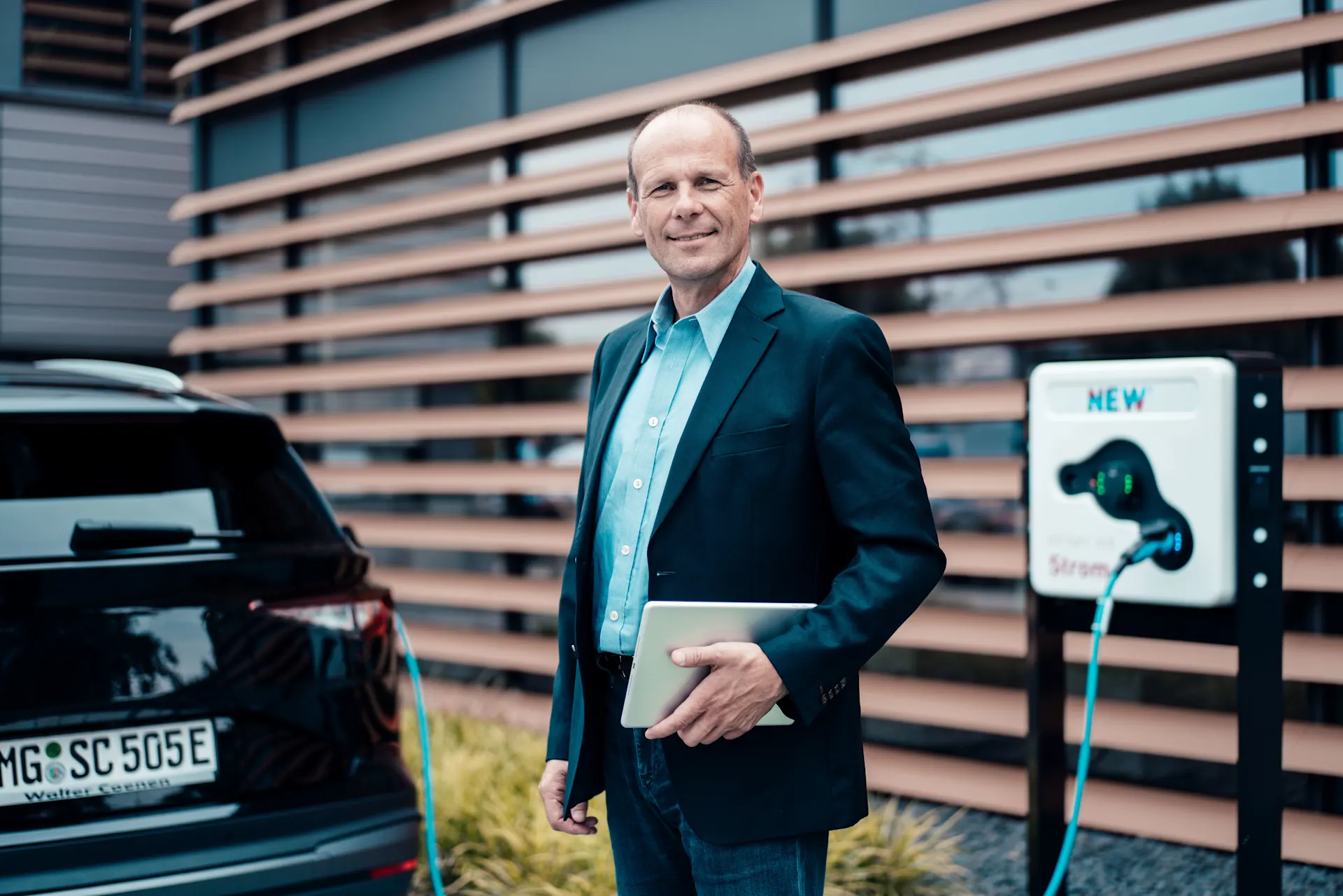 Geschäftsmann steht vor seinem E-Auto mit einem Tablet in der Hand. Das Auto lädt mit Strom der NEW Energie. 