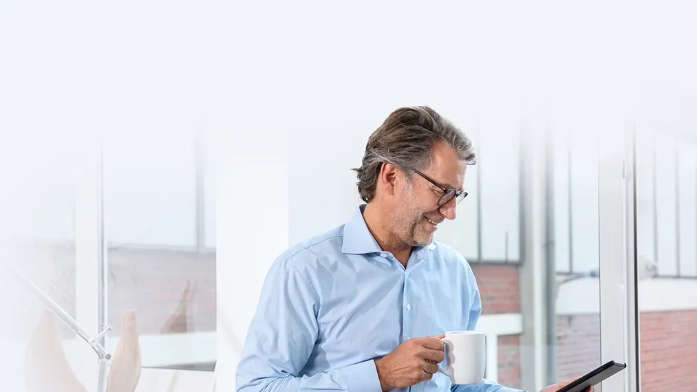 Hausverwalter informiert sich über Serviceangebote der NEW Energie Ein Hausverwalter steht mit einem Kaffee in der Hand vor einem Fenster und schaut mit dem Smartphone nach Serviceangeboten der NEW Energie.