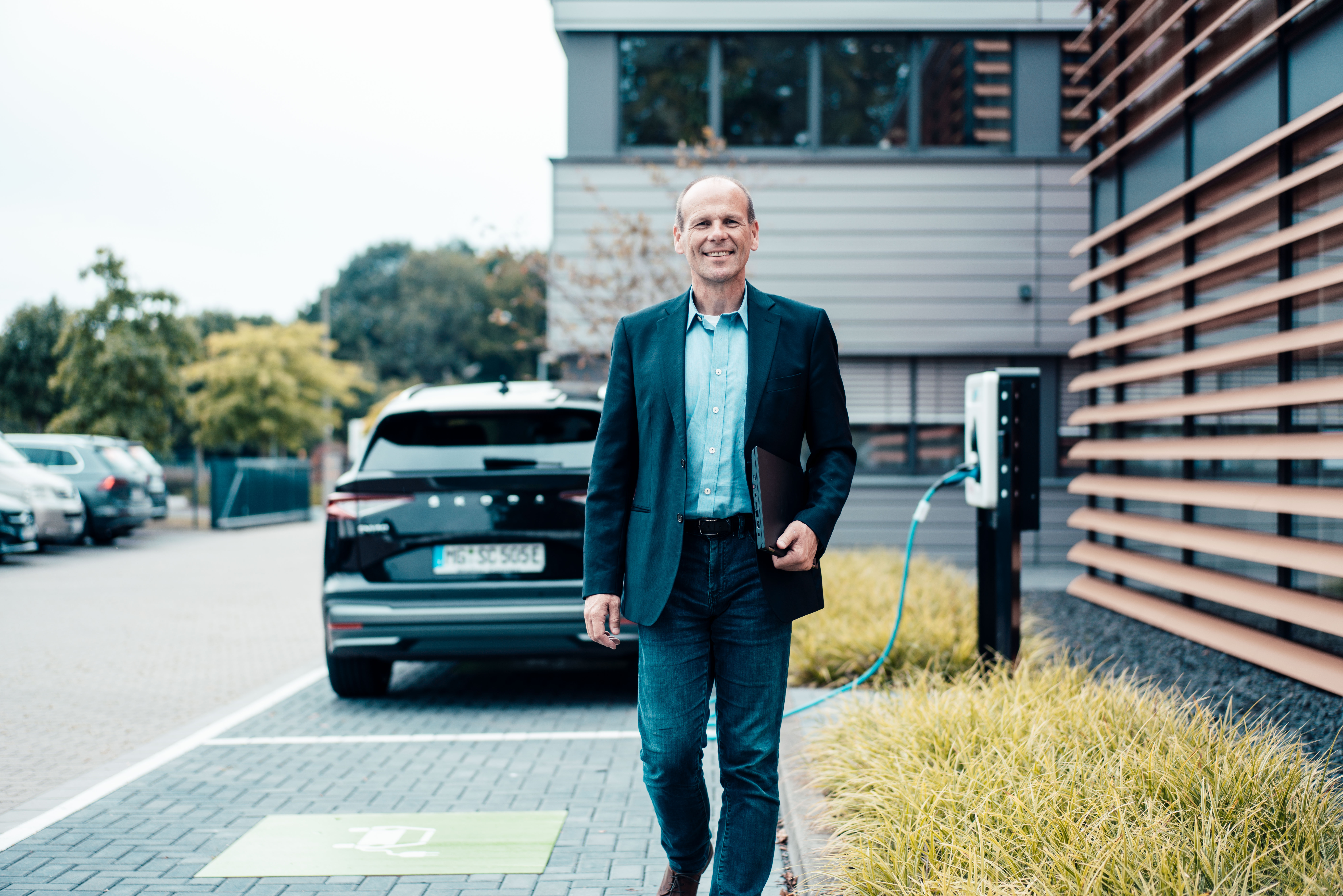 Unternehmer geht von seinem E-Auto weg und hält einen Laptop in der Hand, was mithilfe einer Wallbox auflädt.